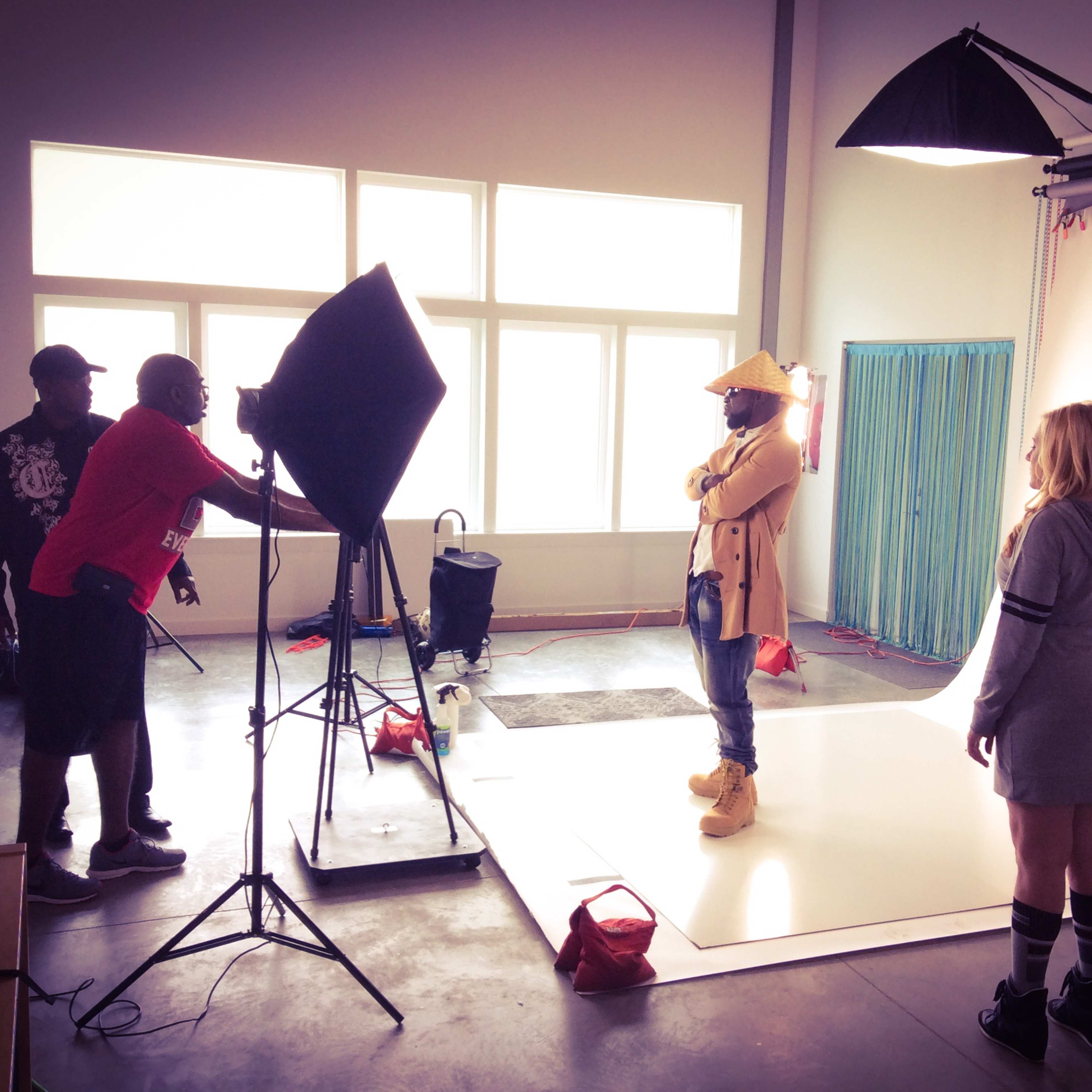 A photoshoot is taking place in a studio, featuring a model in the center with three crew members adjusting equipment and lighting.