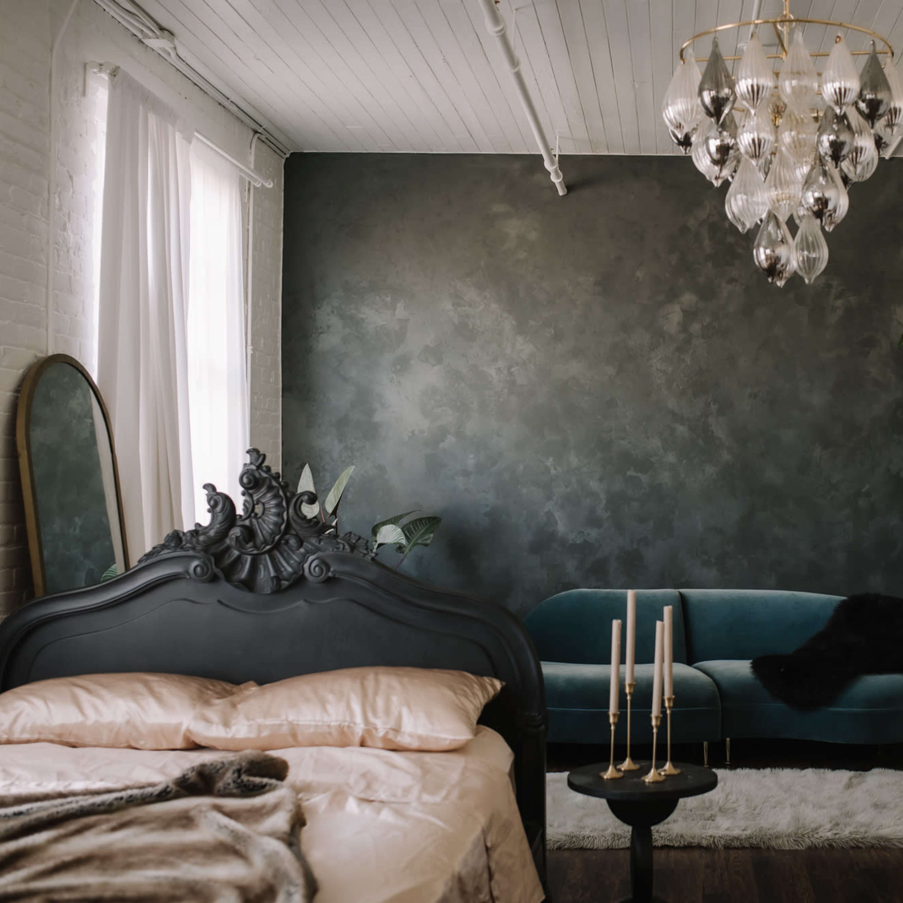 A bedroom features a black ornate bed, a teal sofa against a dark green wall, and a floor plant, with candles on a small table.