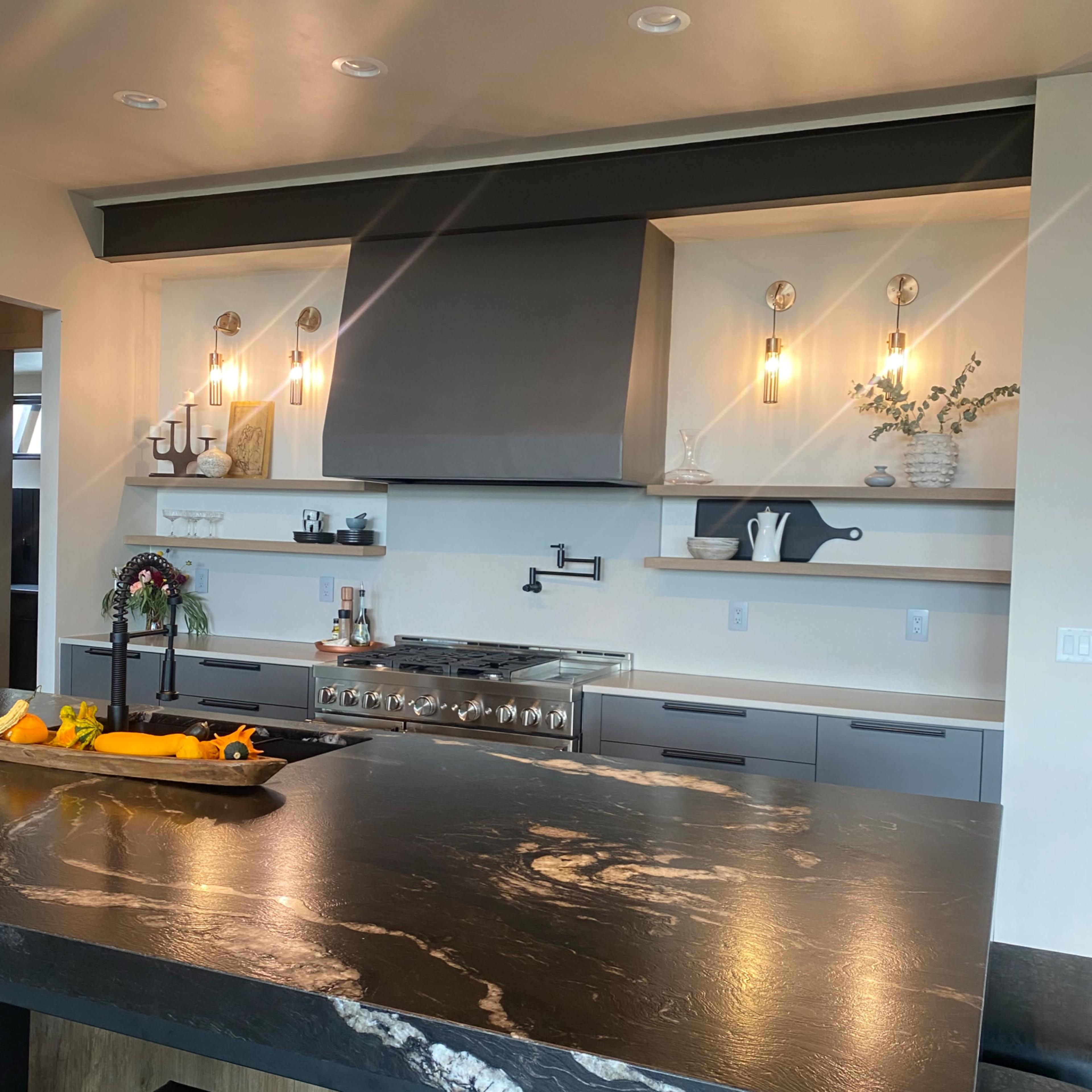 A modern kitchen featuring a large dark stone island, a stainless steel stove with a hood above, and minimalist shelving with decorative items.