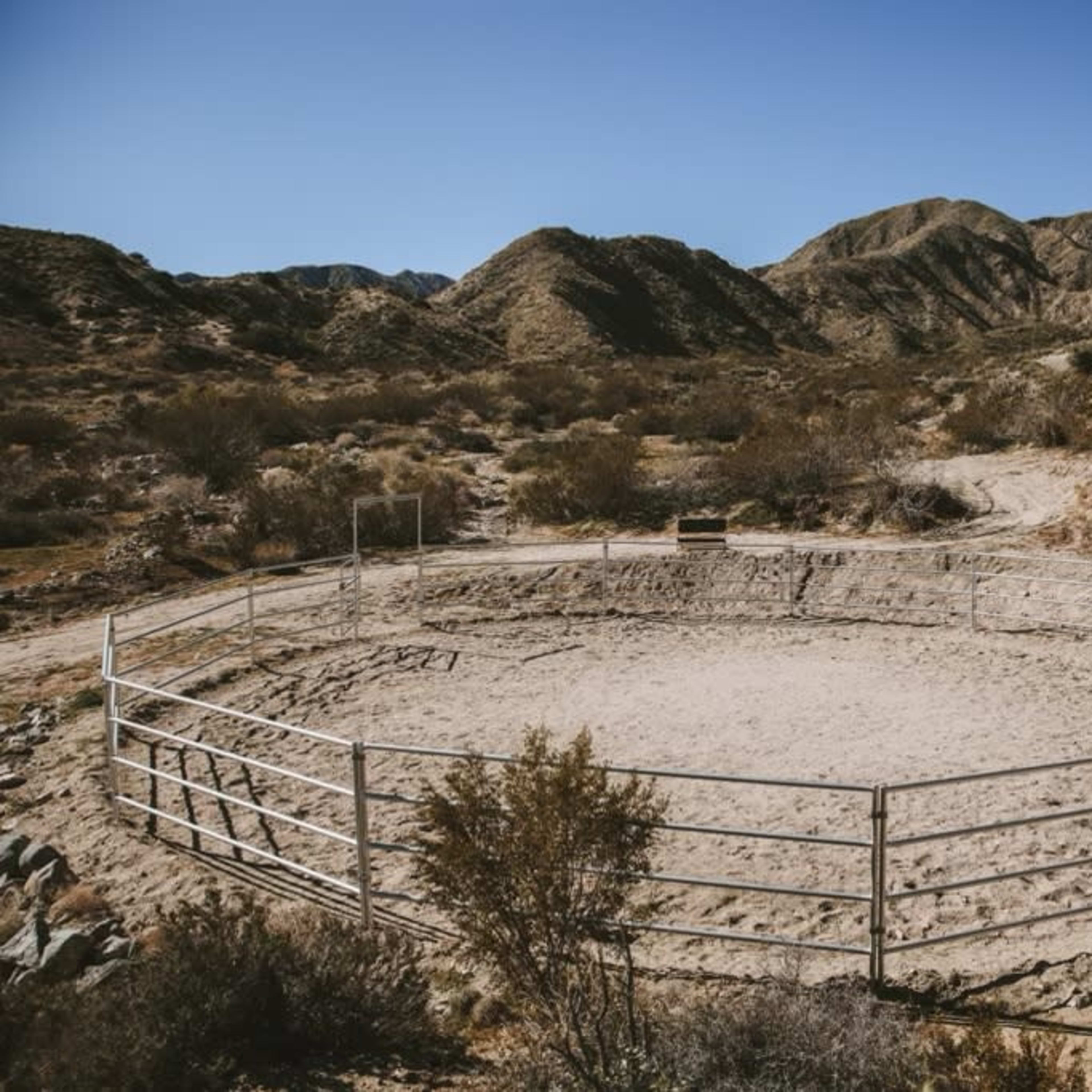 A circular dirt arena enclosed by a metal fence is situated among dry, hilly terrain.