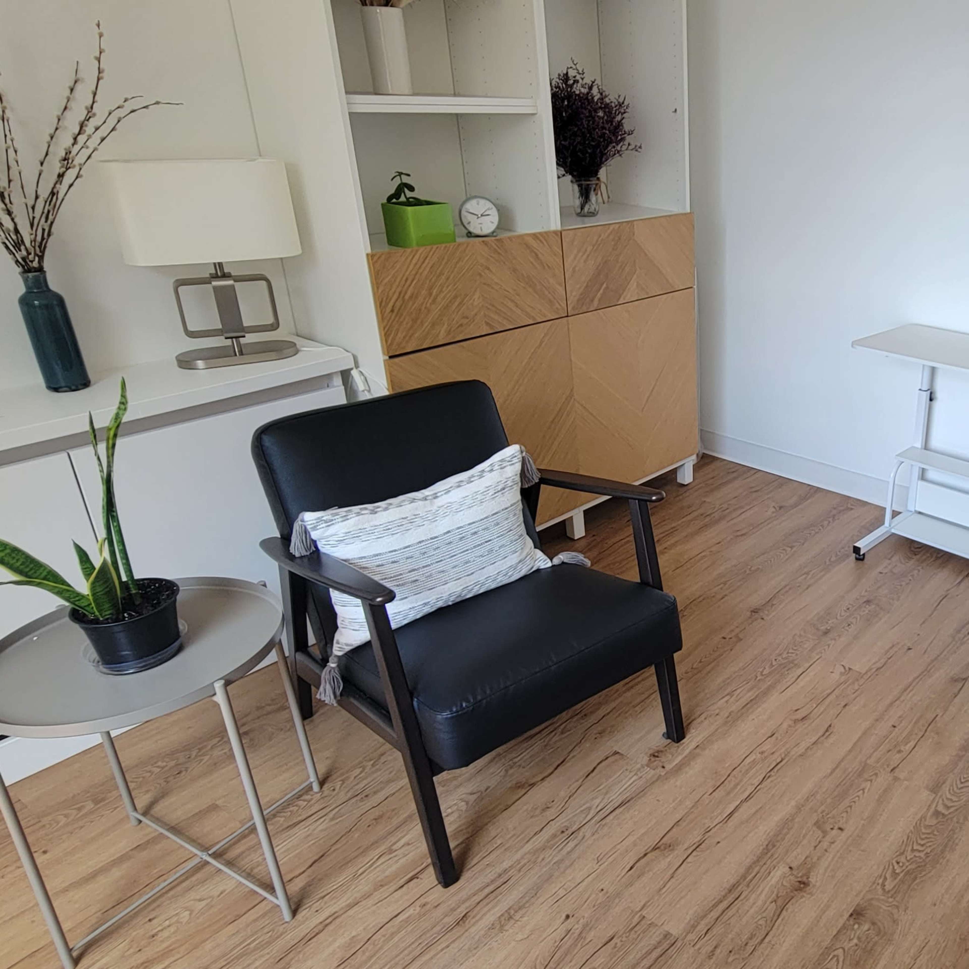 A neat room featuring a black chair, a small round table, a white folding table, and a gray sofa against light-colored walls and wooden flooring.