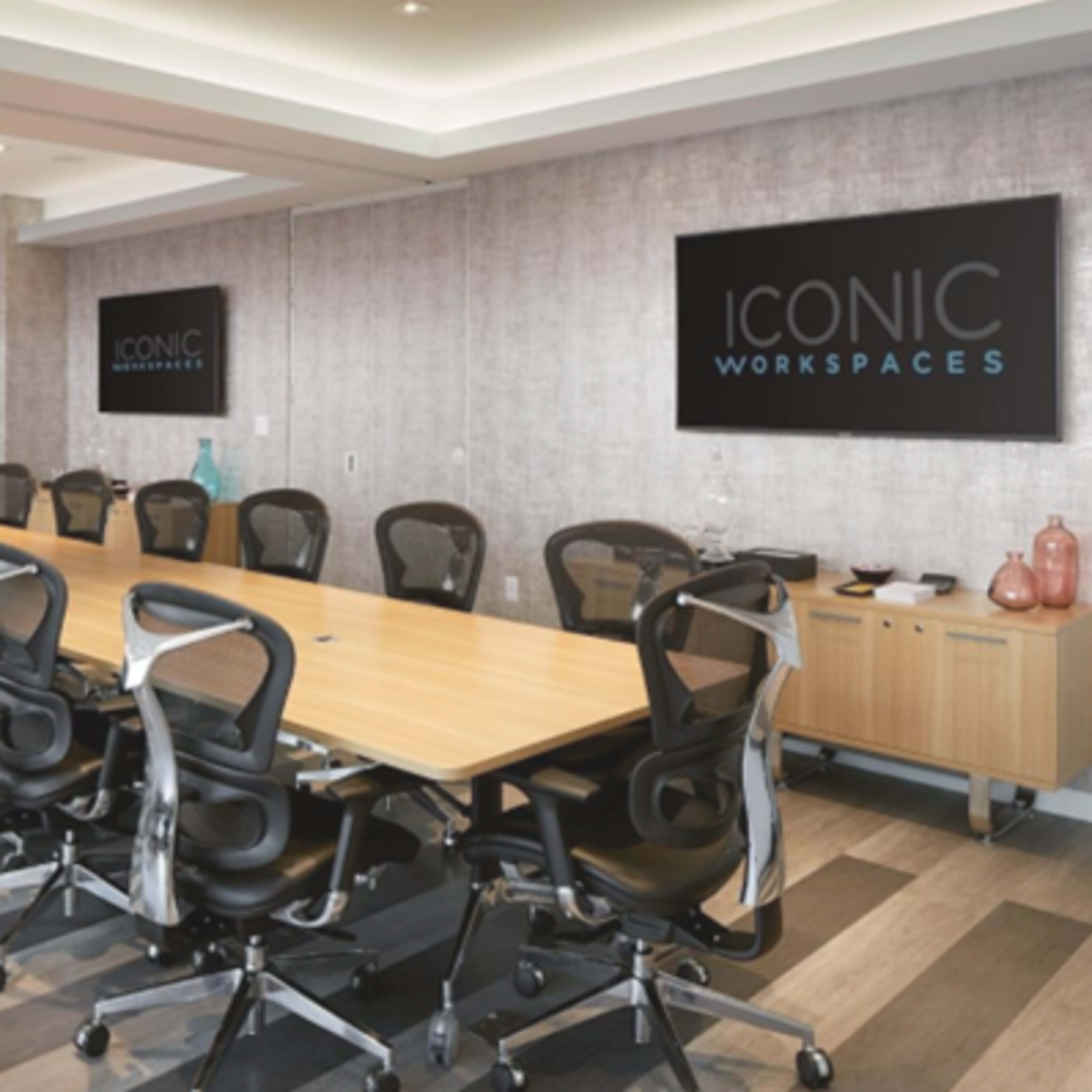 A modern conference room features a long wooden table surrounded by ergonomic black chairs, with large screens displaying the text "ICONIC WORKSPACES" on the walls.