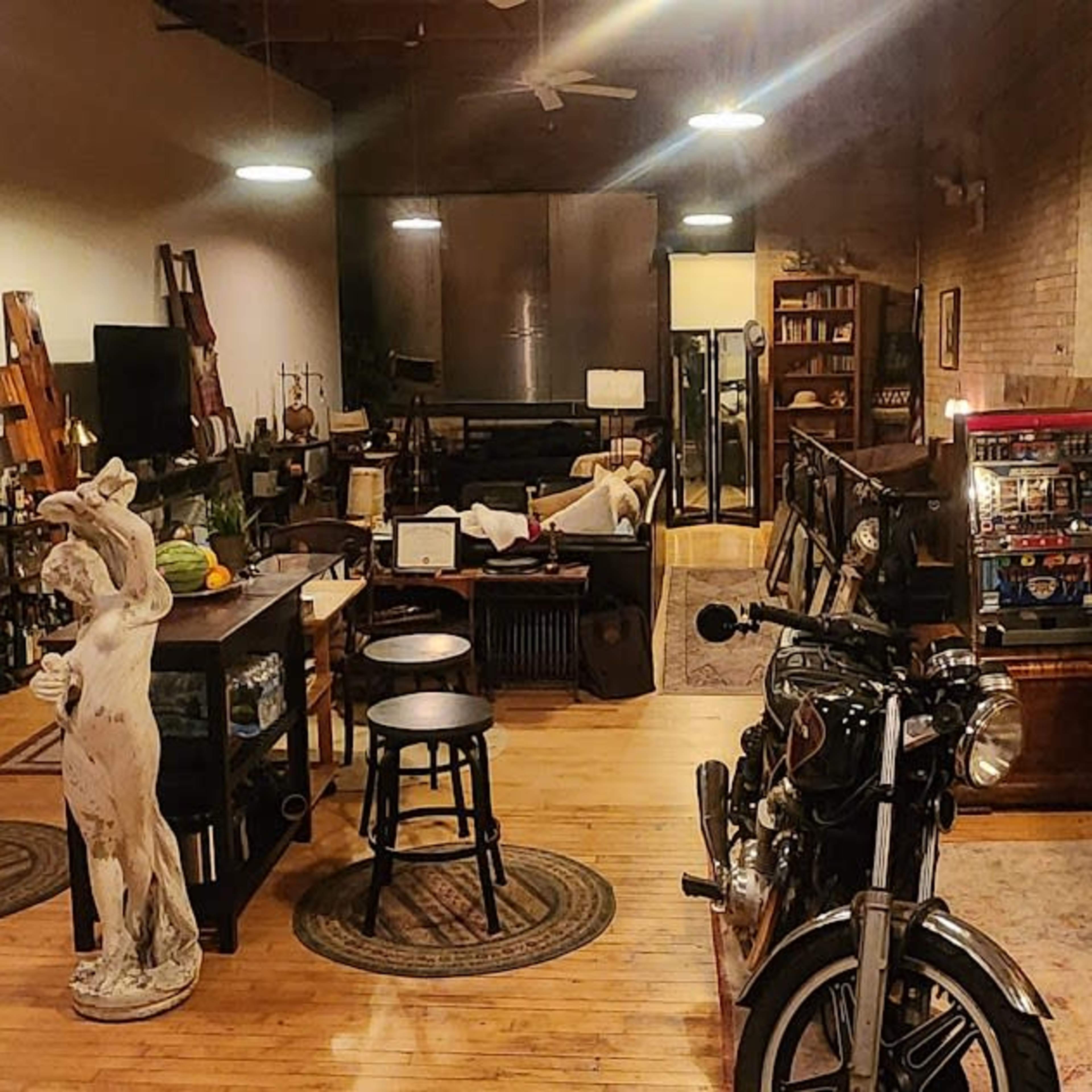A spacious industrial-style loft featuring a kitchen area, a motorcycle, a vintage pinball machine, and various pieces of furniture.