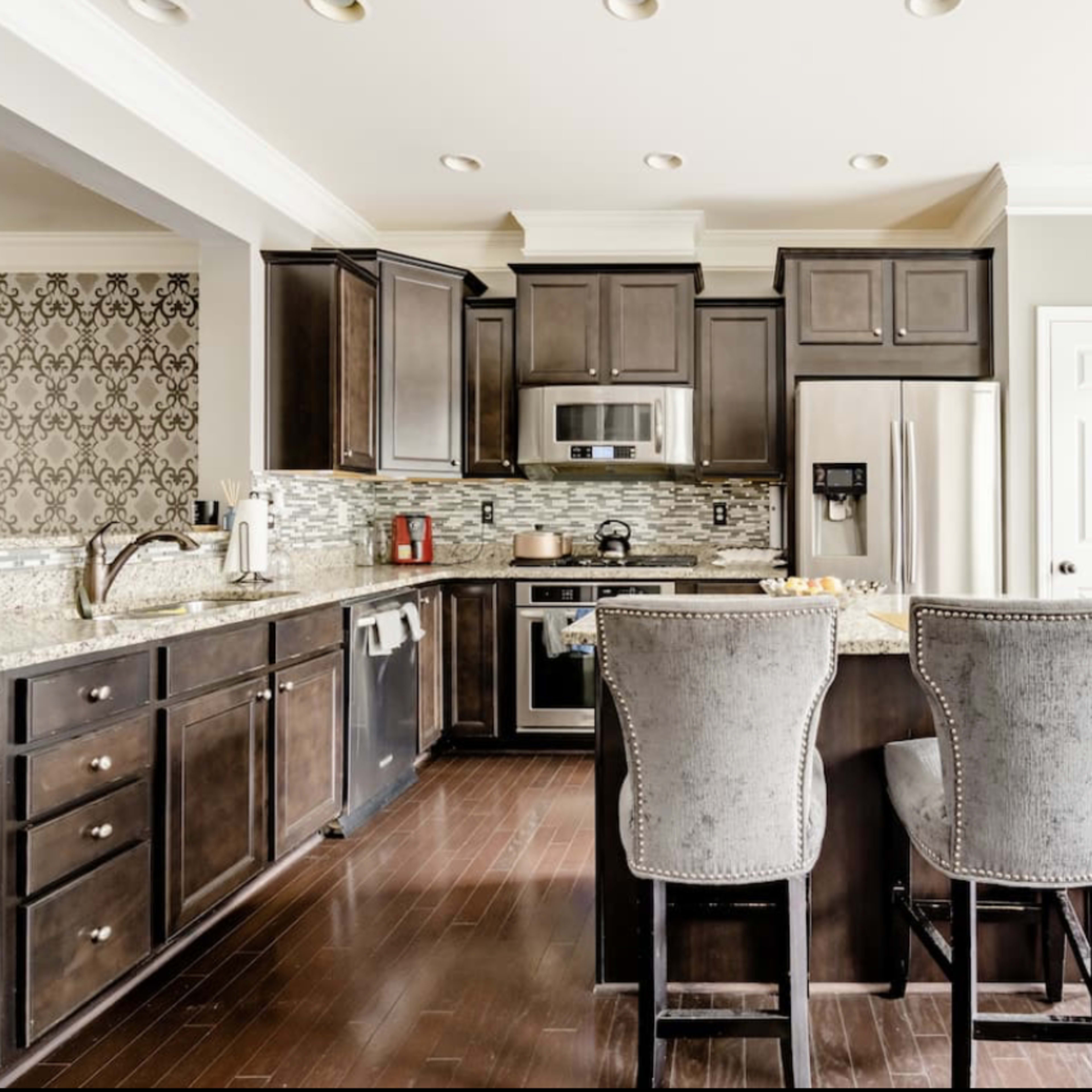 A modern kitchen featuring dark wooden cabinets, stainless steel appliances, a granite countertop, and two upholstered bar stools.