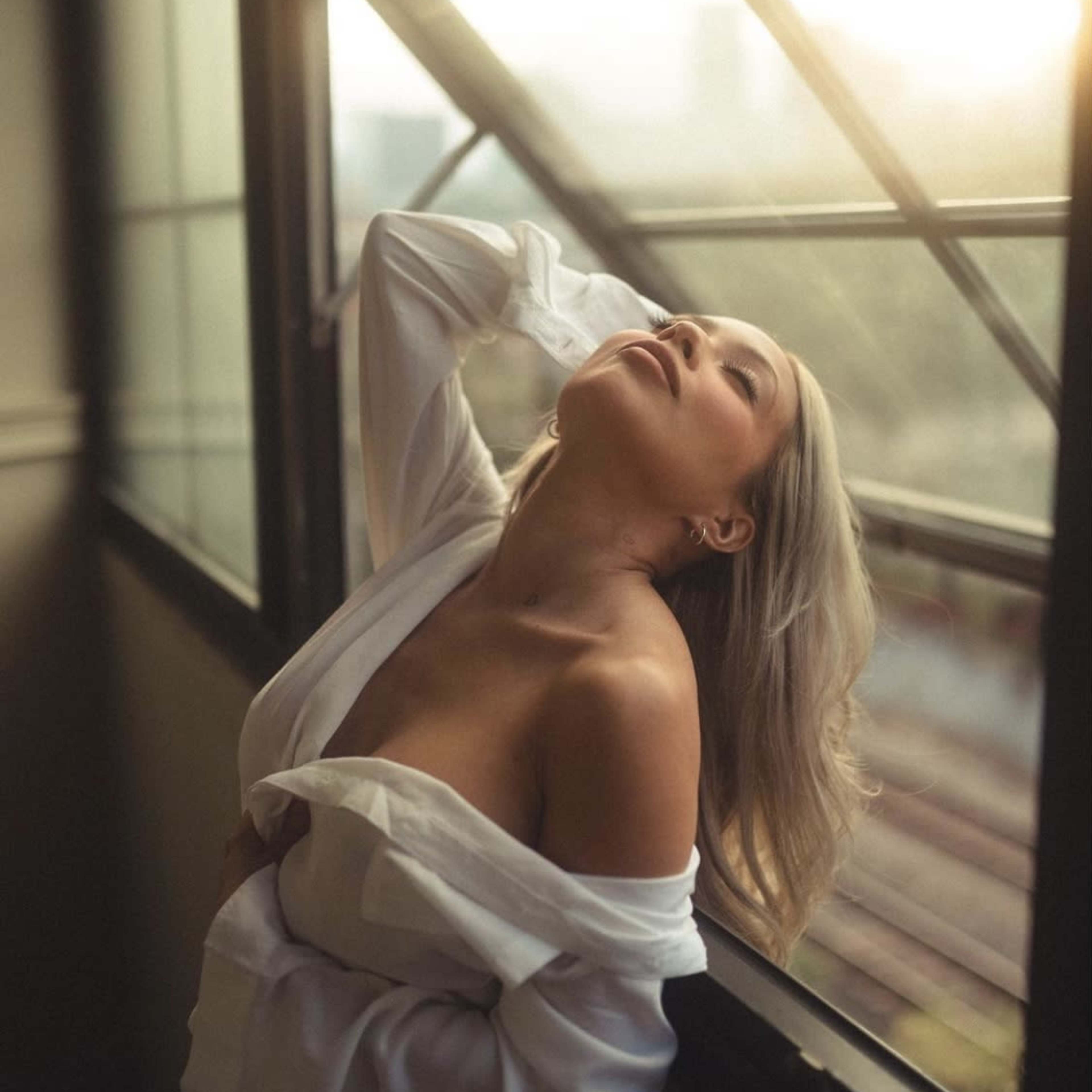 A person in a white shirt leans against a window, with sunlight streaming in from the outside.