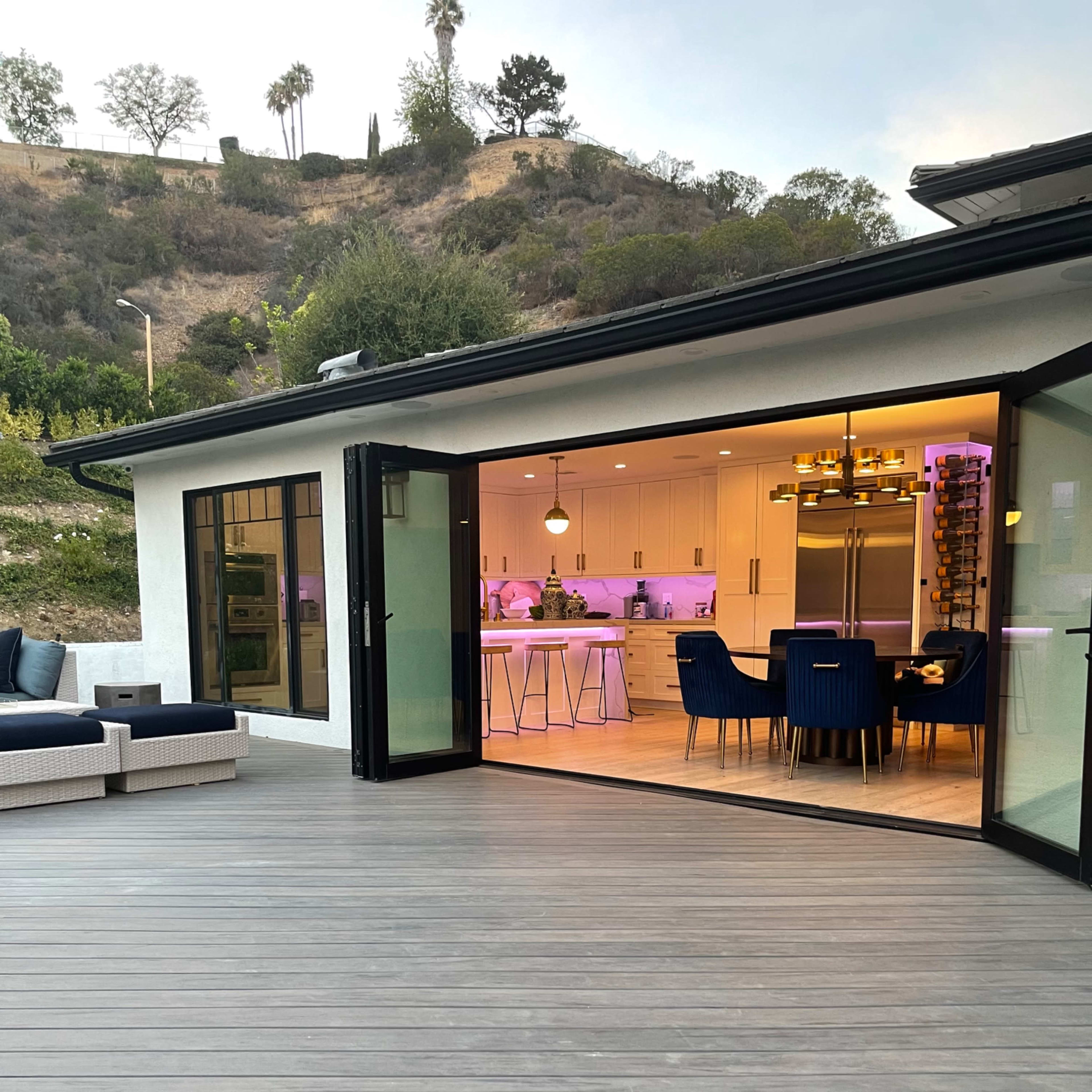 A modern home with large glass doors leading to a deck, overlooking a hillside with trees, and an interior kitchen area illuminated with colorful lighting.