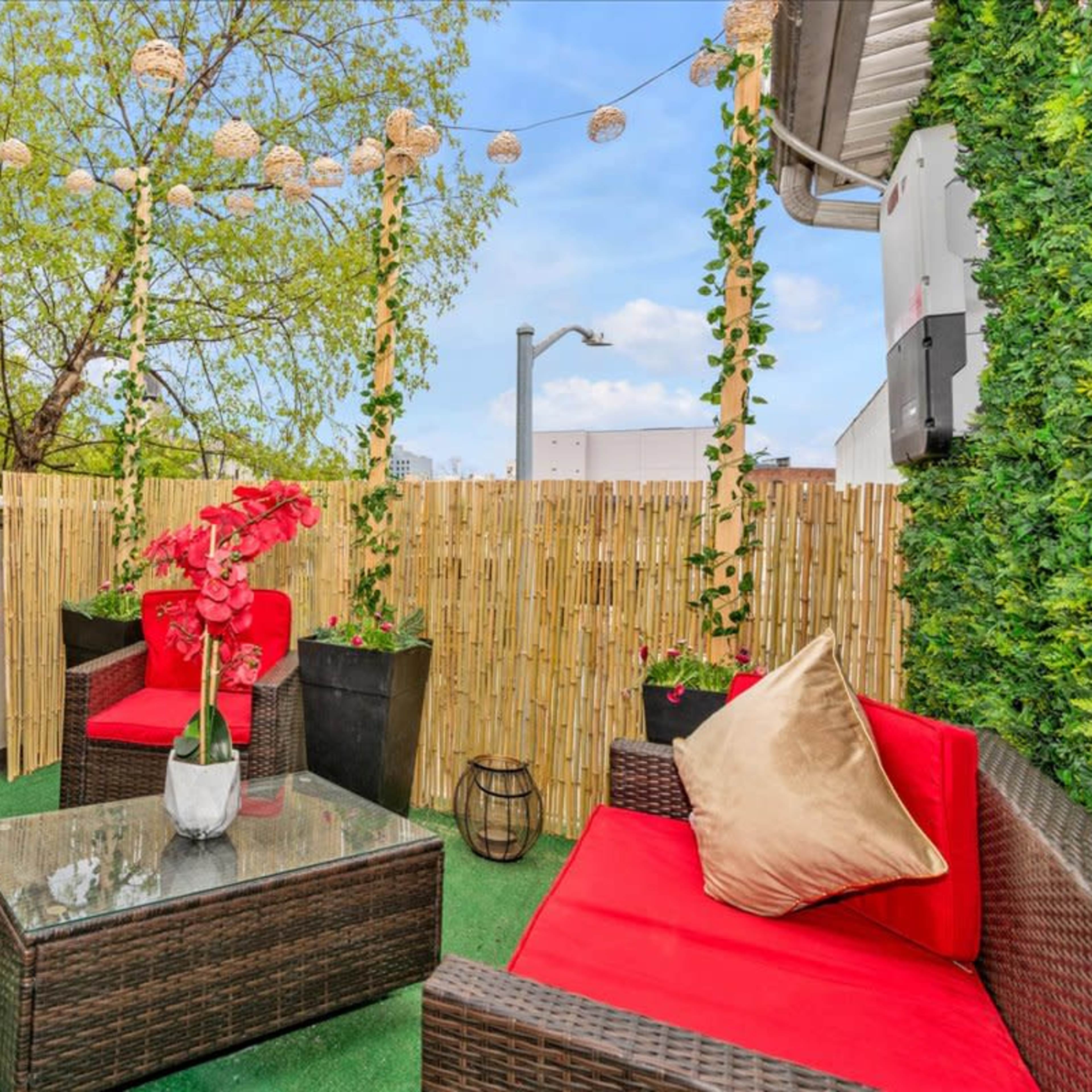 A rooftop terrace with red outdoor furniture, potted plants, and decorative lighting against a backdrop of greenery.