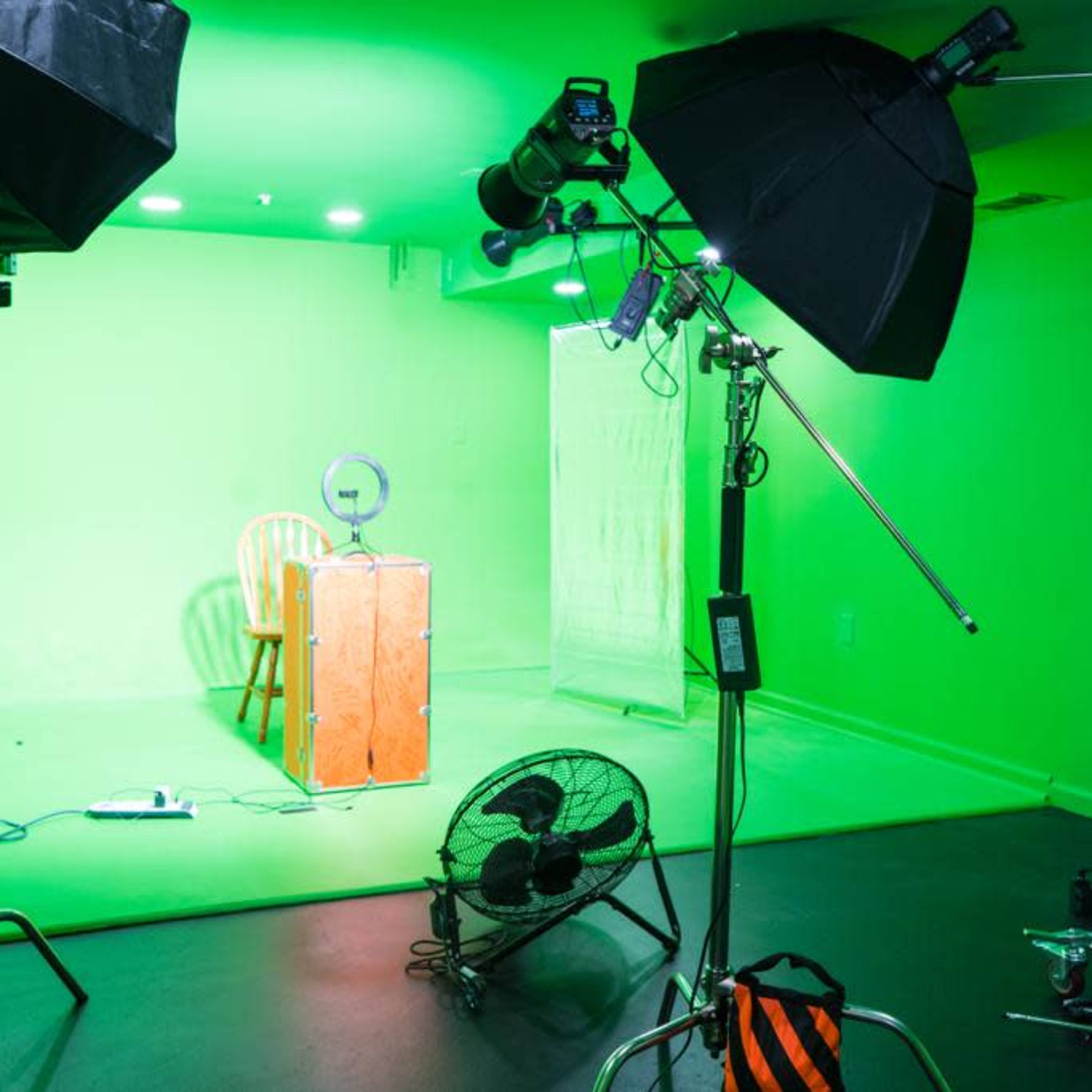 A green screen studio setup with lighting equipment, a chair, a table, and a fan.