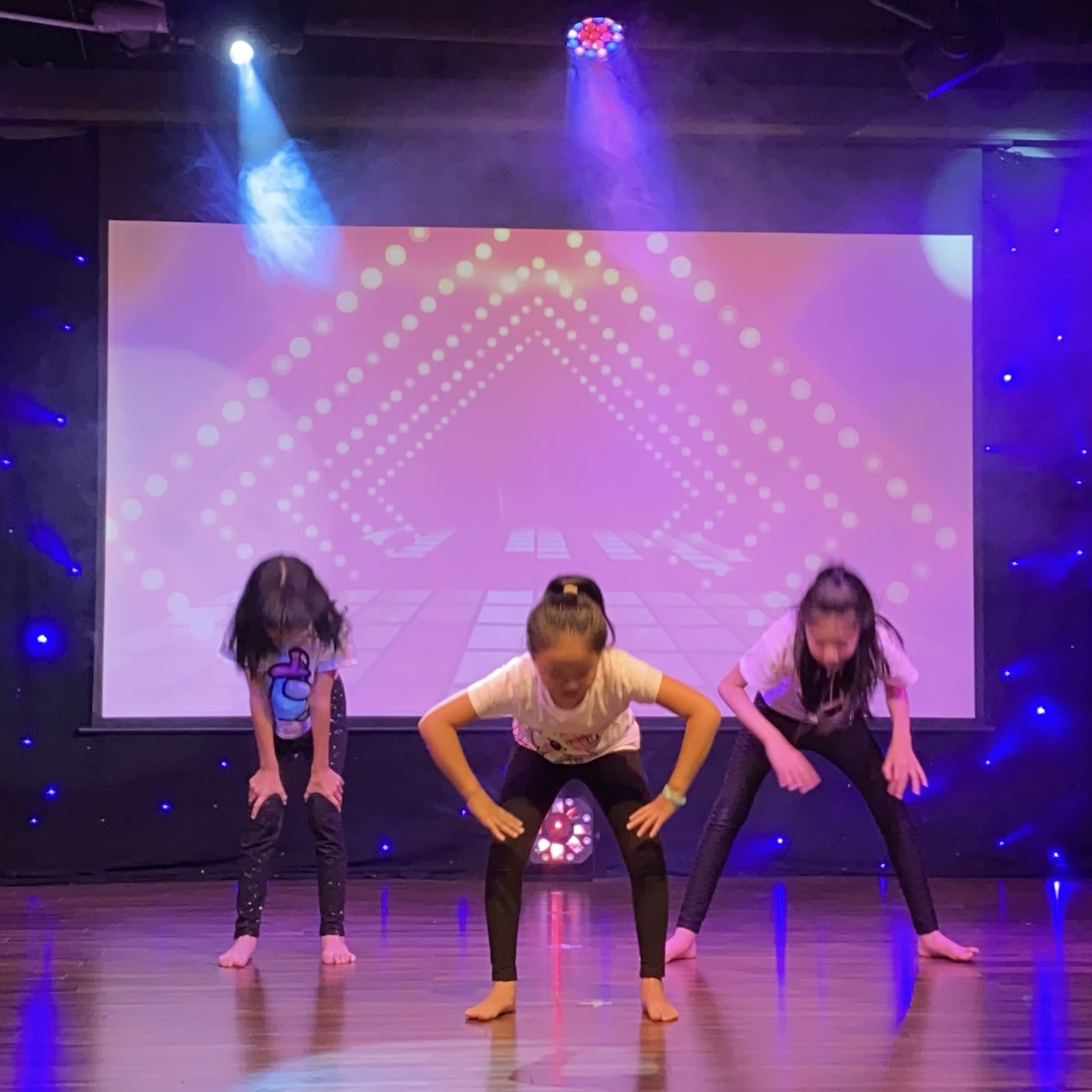 Three children perform a dance routine on stage with a colorful backdrop and stage lights.