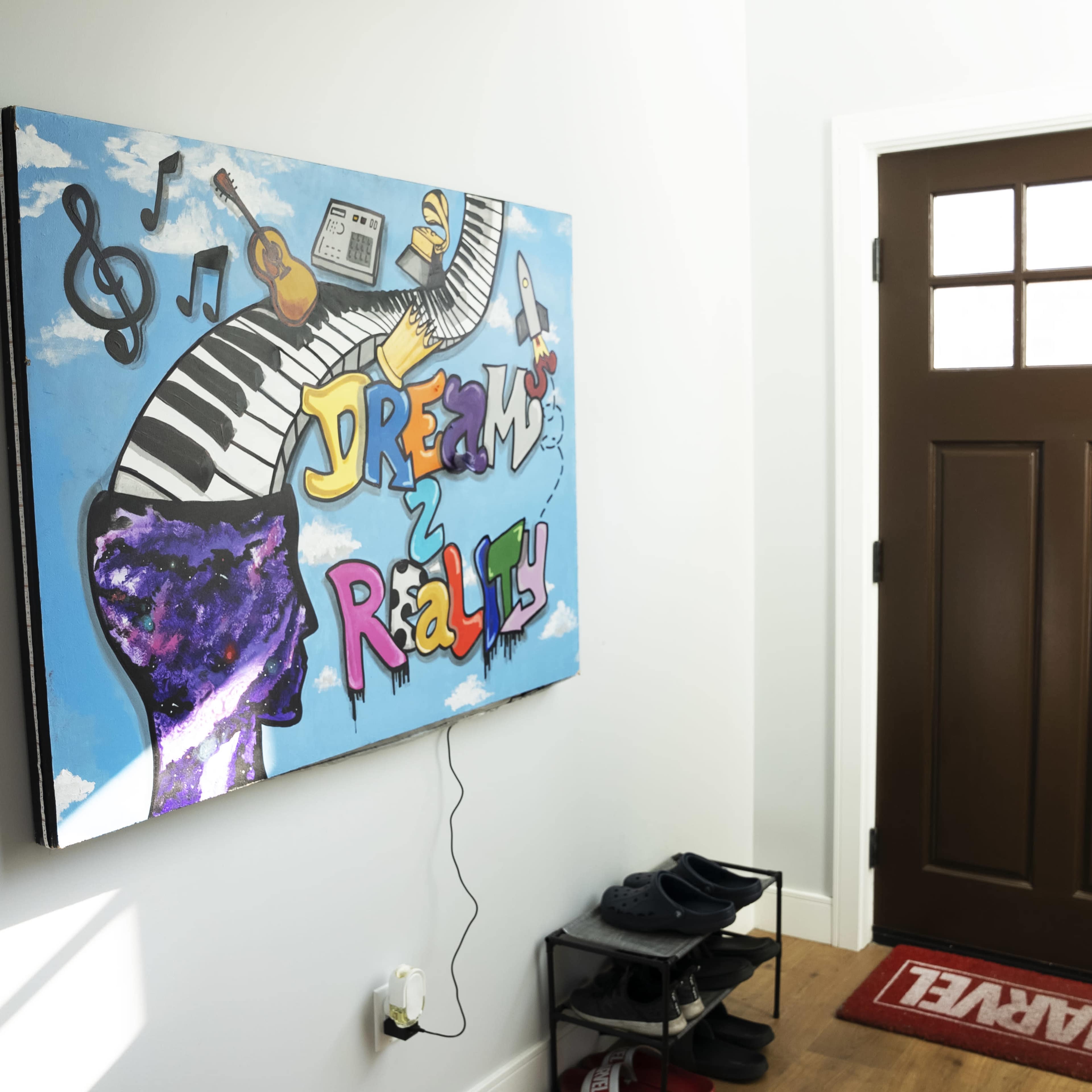 A colorful mural titled "Dreams 2 Reality" hanging on a wall near a brown front door with a window and a red doormat featuring the Marvel logo.