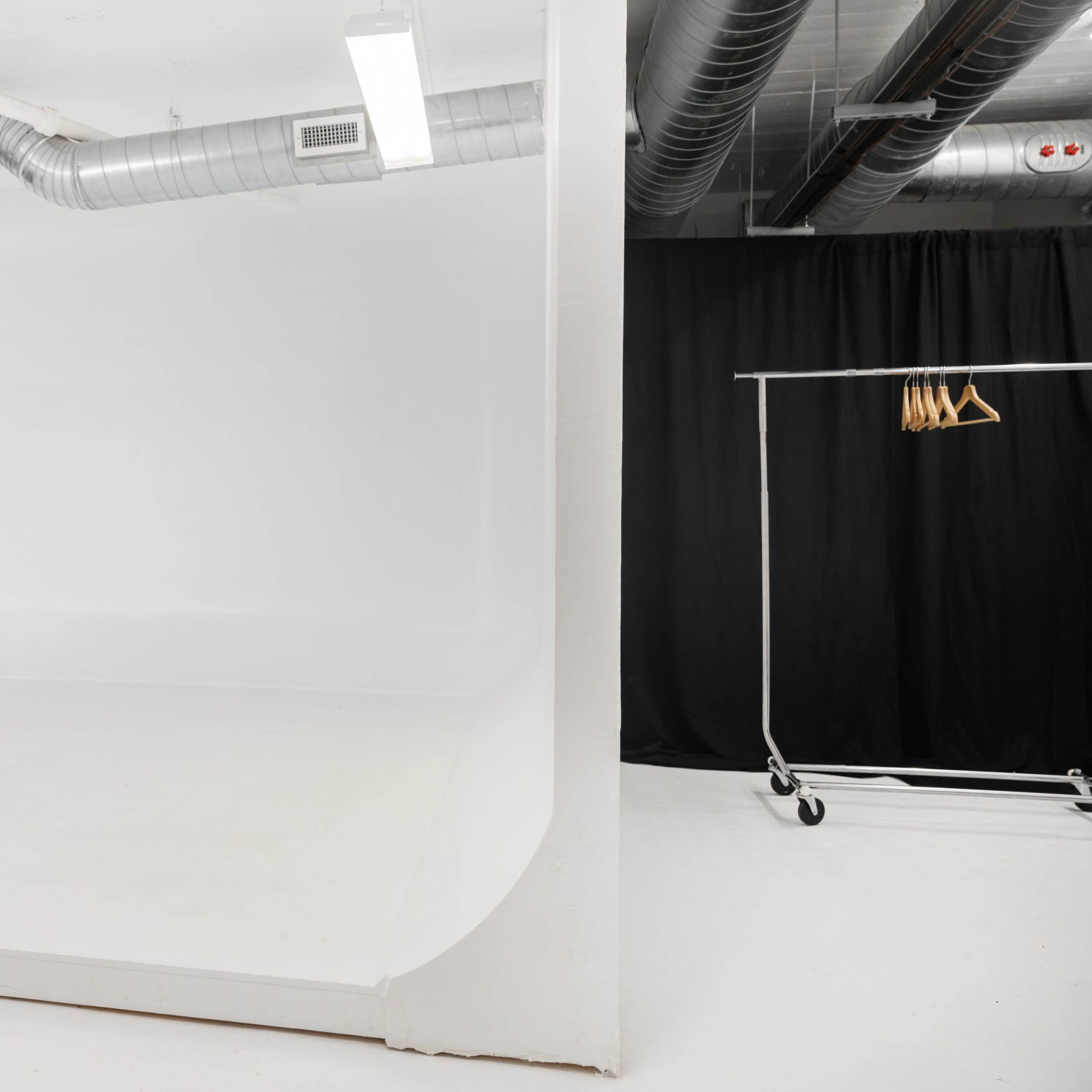 A photography studio with a large white backdrop and a clothing rack on wheels with hangers against a black curtain.