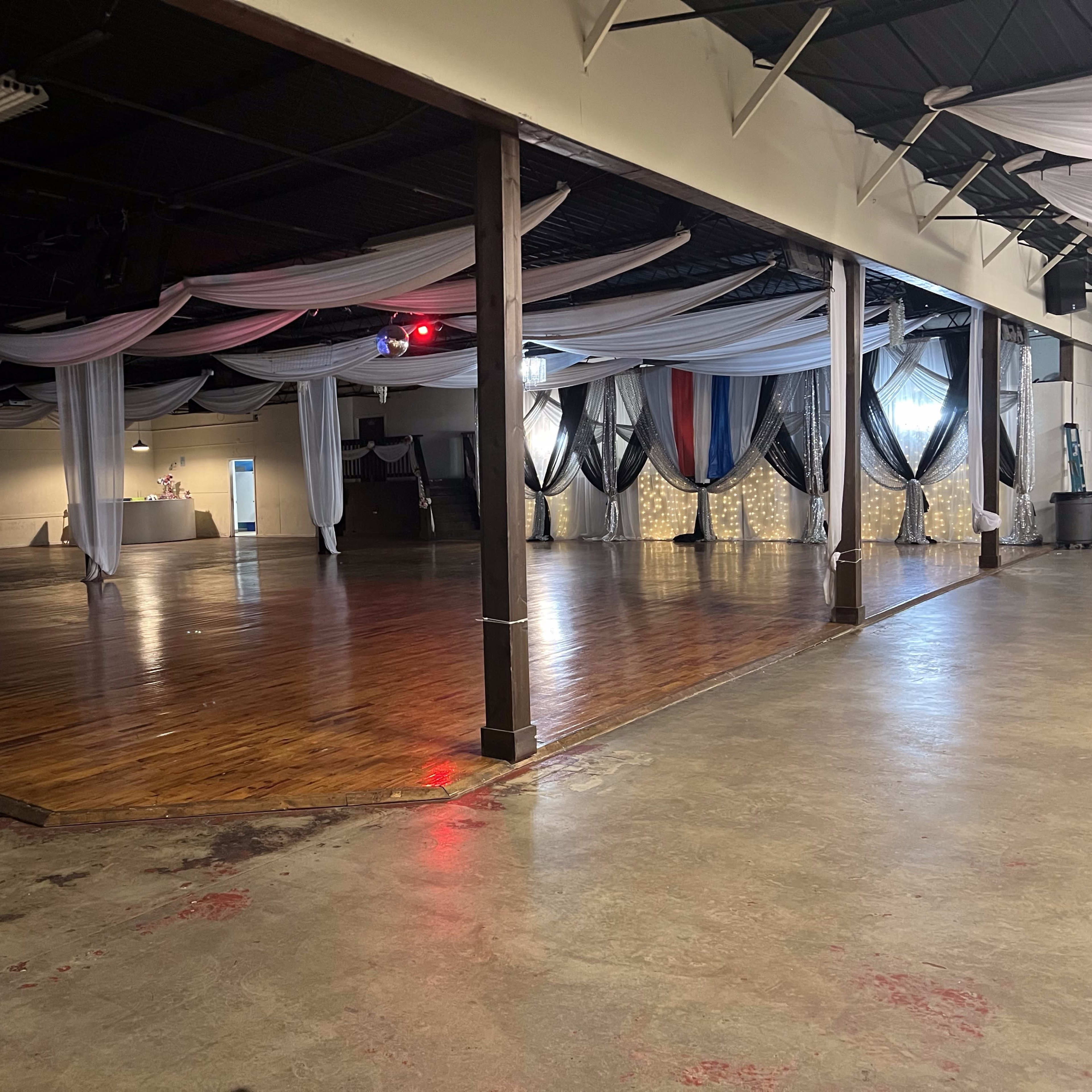 A spacious indoor venue with wooden flooring, draped fabric hanging from the ceiling, and a projection screen displaying an image on one wall.