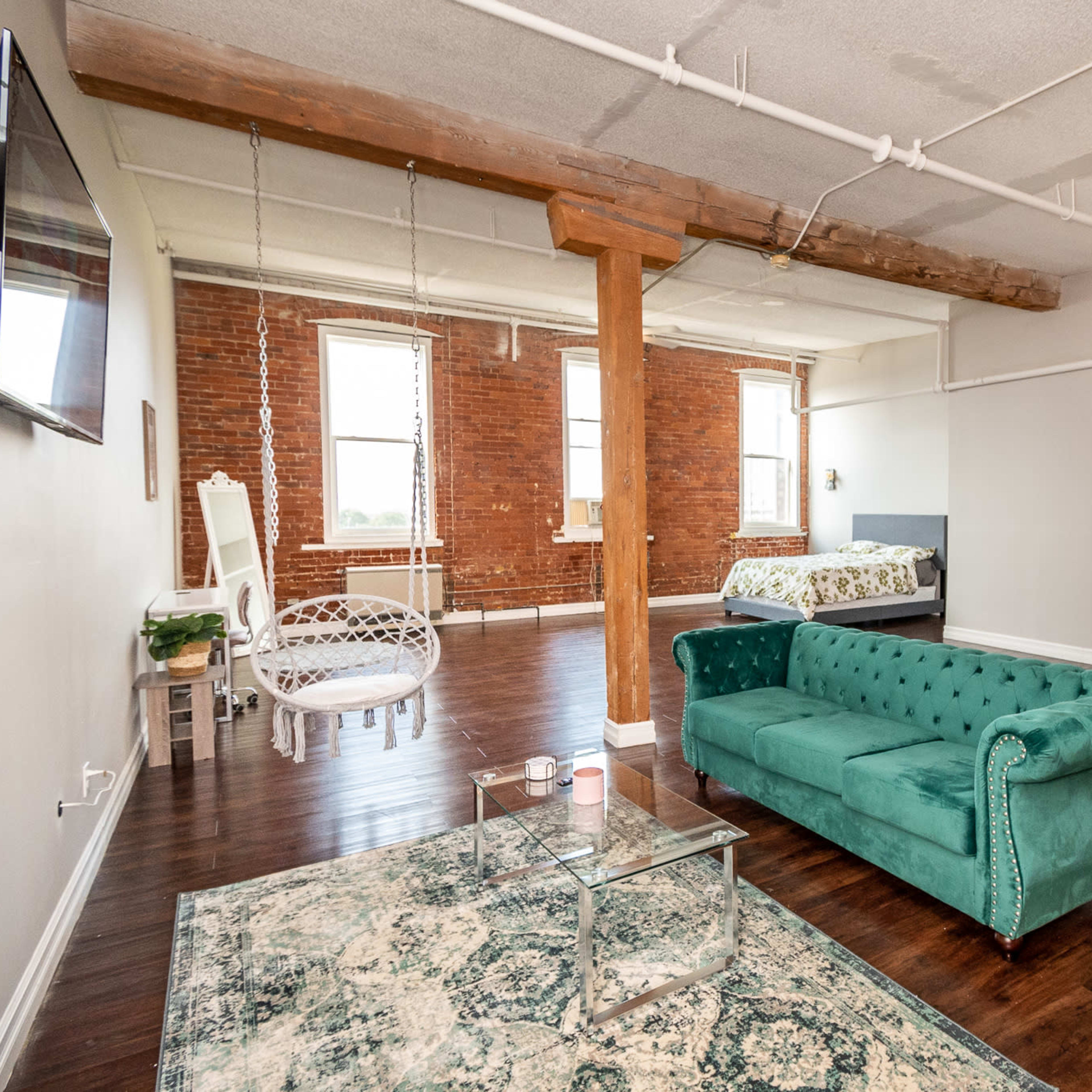 A spacious, modern living area with a green velvet sofa, a glass table with chairs, and exposed brick walls featuring large windows.