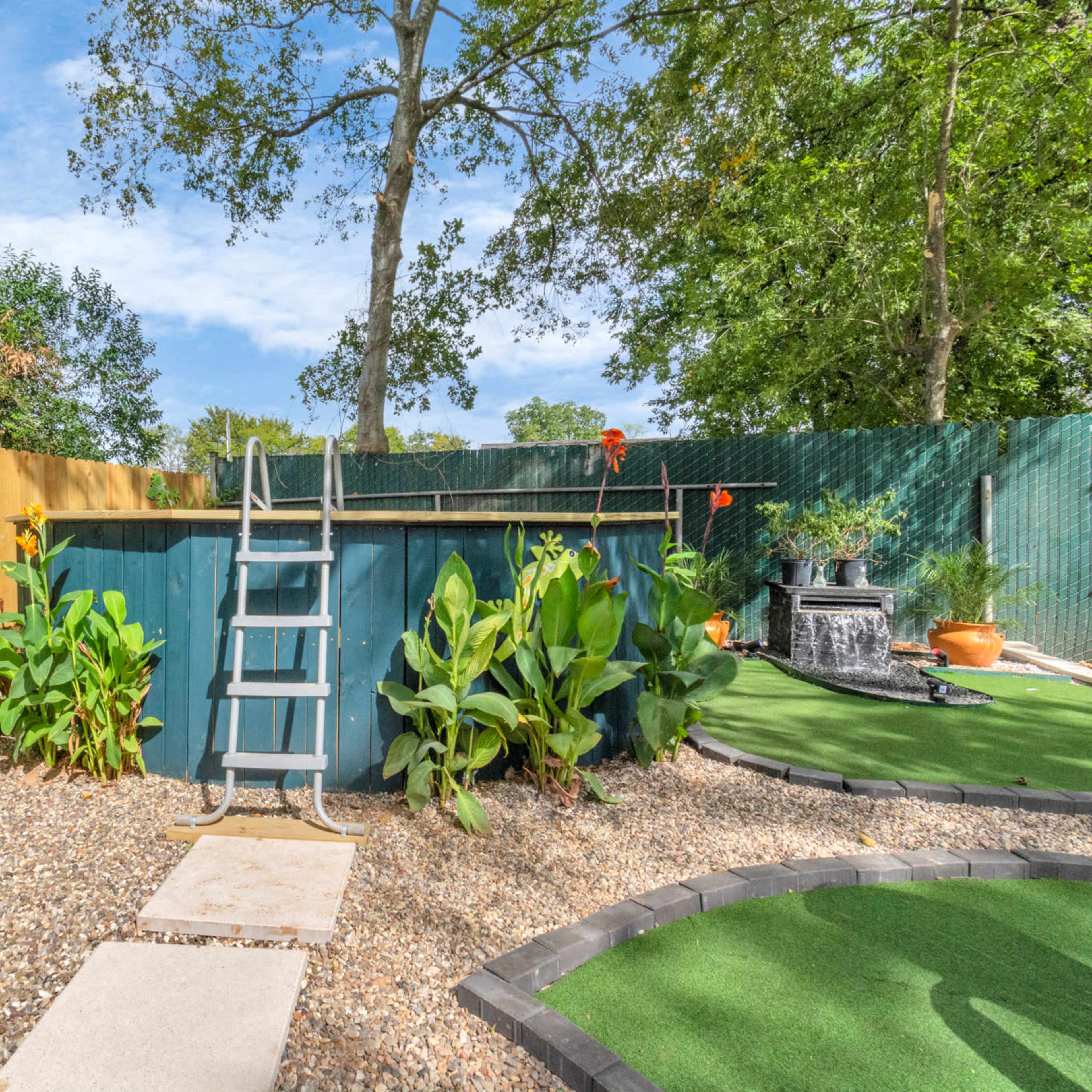 A small, above-ground swimming pool with a ladder is surrounded by landscaped greenery and a gravel path in a backyard setting.