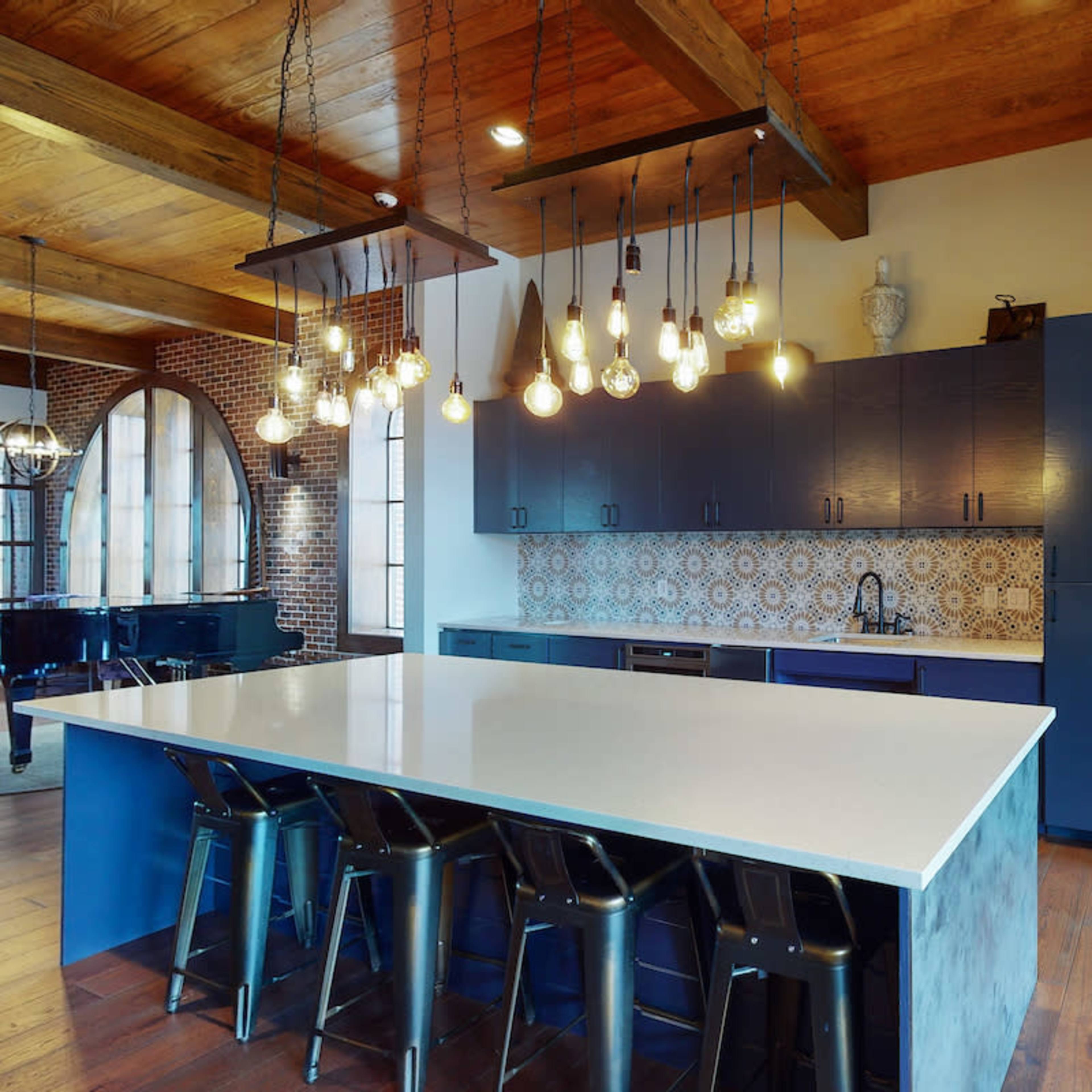 A modern kitchen features a large island with seating, dark cabinetry, and pendant lights above, set against a backdrop of exposed brick and wooden beams.