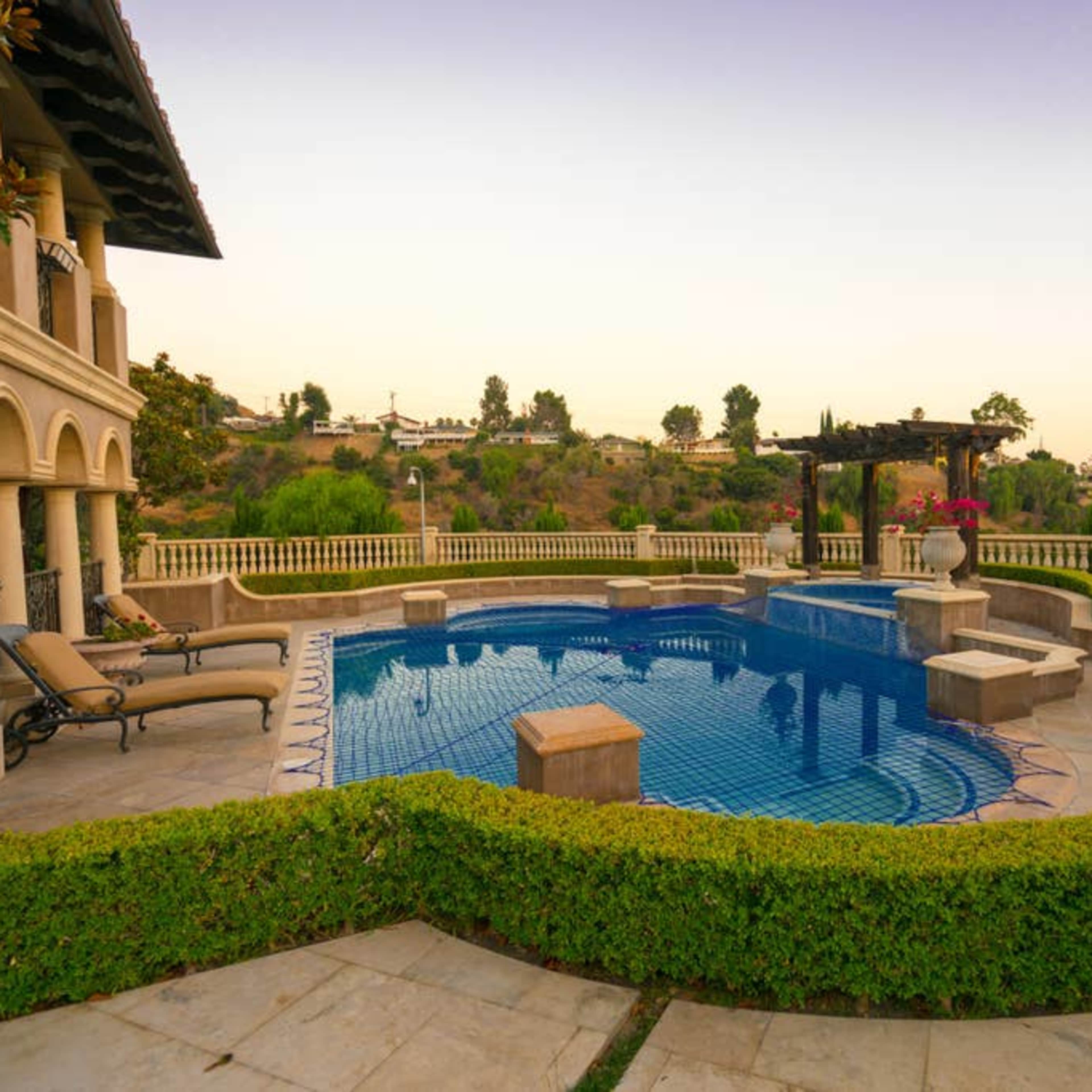 A luxurious pool area surrounded by manicured hedges and a hillside landscape at sunset.