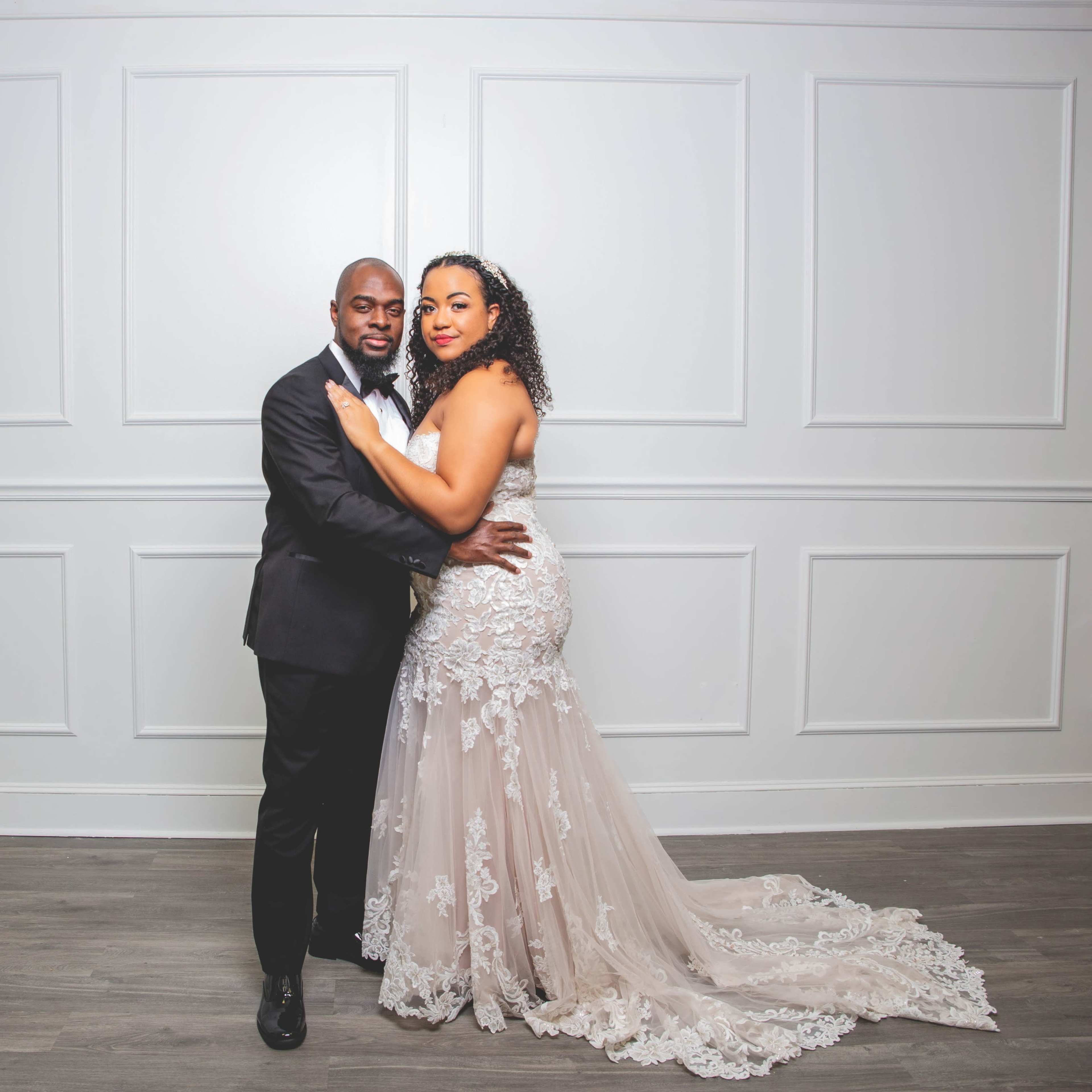 A man in a black tuxedo and a woman in a white wedding dress embrace in front of a plain white wall.