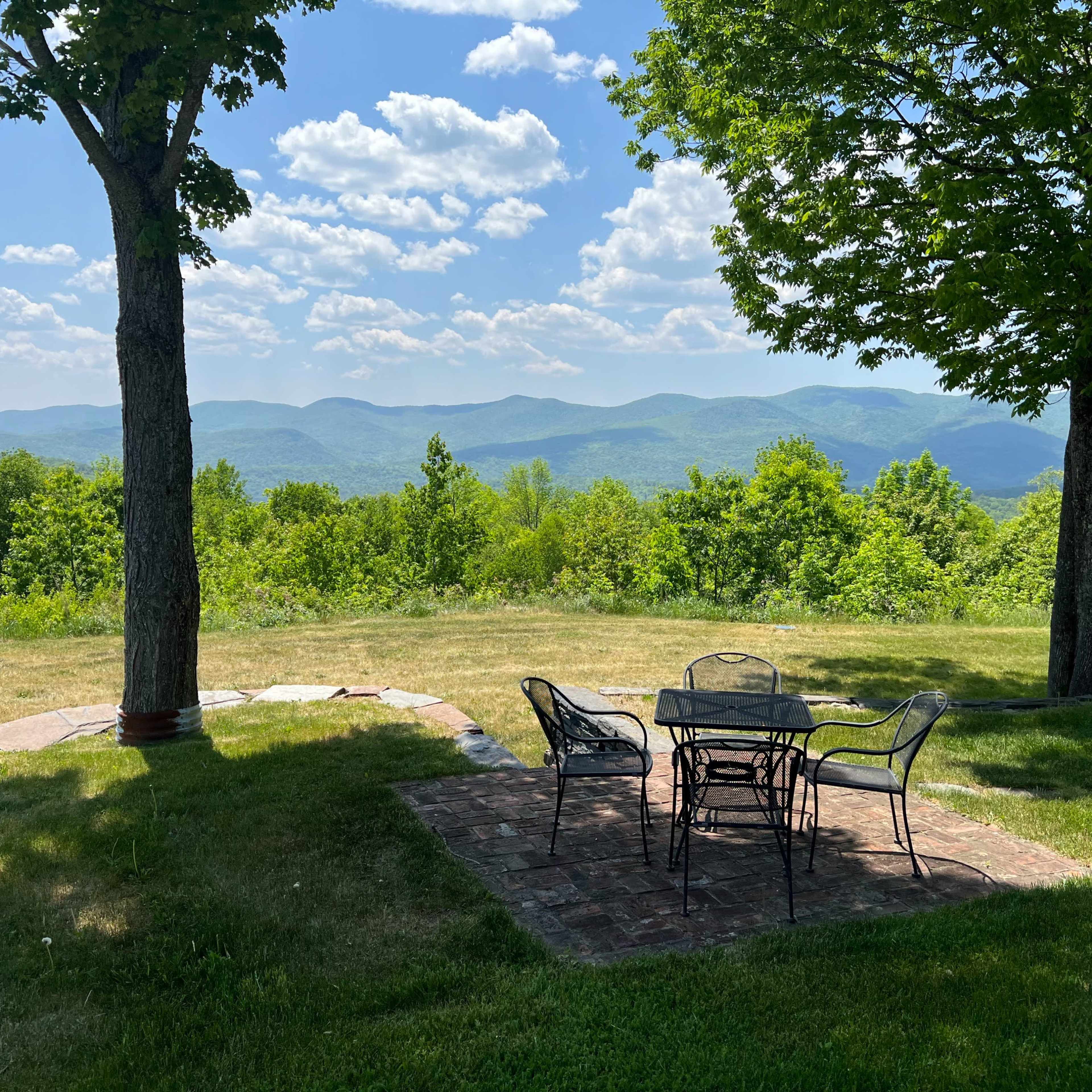 A stone patio set with a table and chairs overlooks a valley of rolling green hills and distant mountains under a partly cloudy sky.