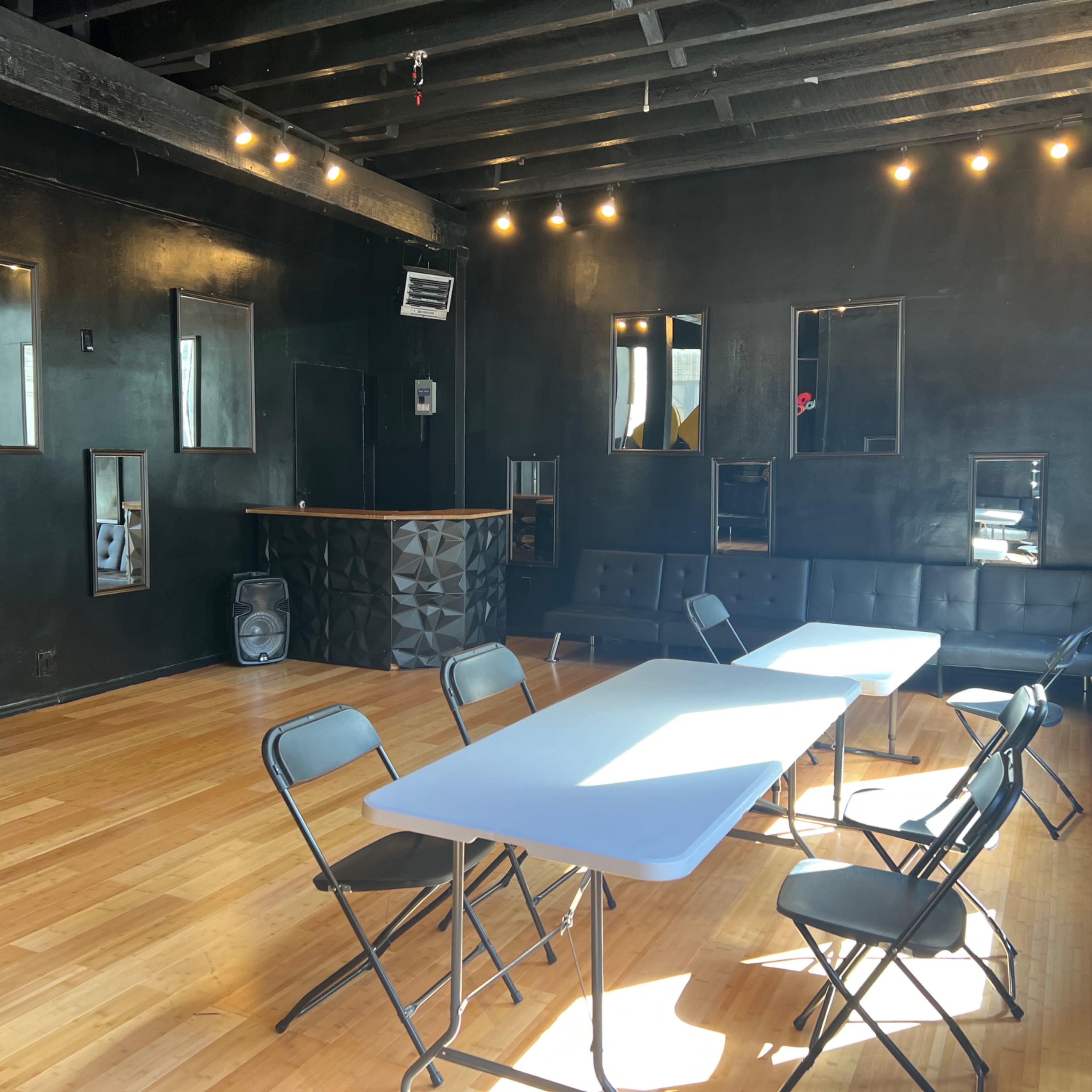 A spacious room with black walls, wooden flooring, a bar counter, and several tables and chairs arranged for an event.