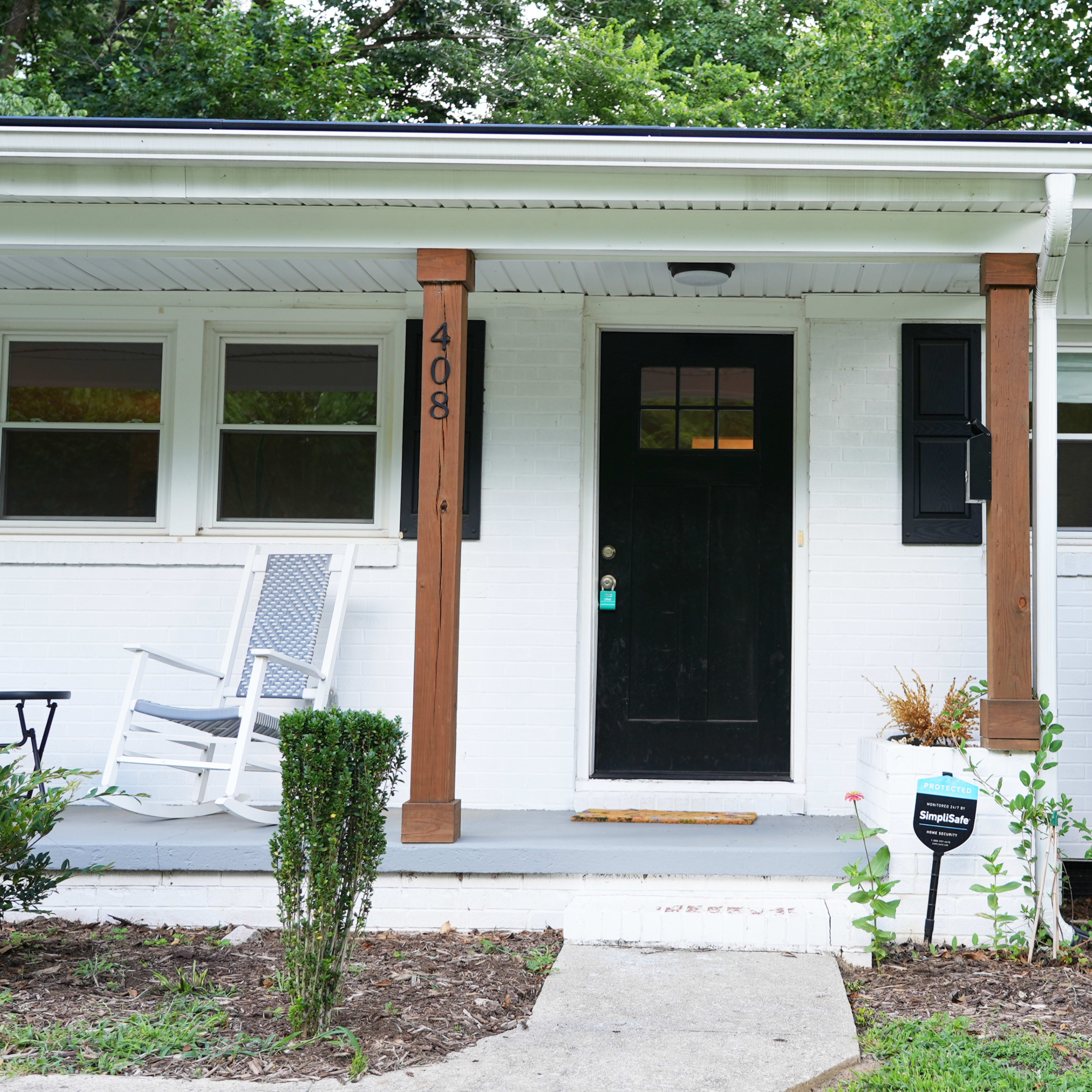 A white house with a black front door, wooden columns, a porch with a rocking chair, and neatly trimmed bushes flanking the entrance.