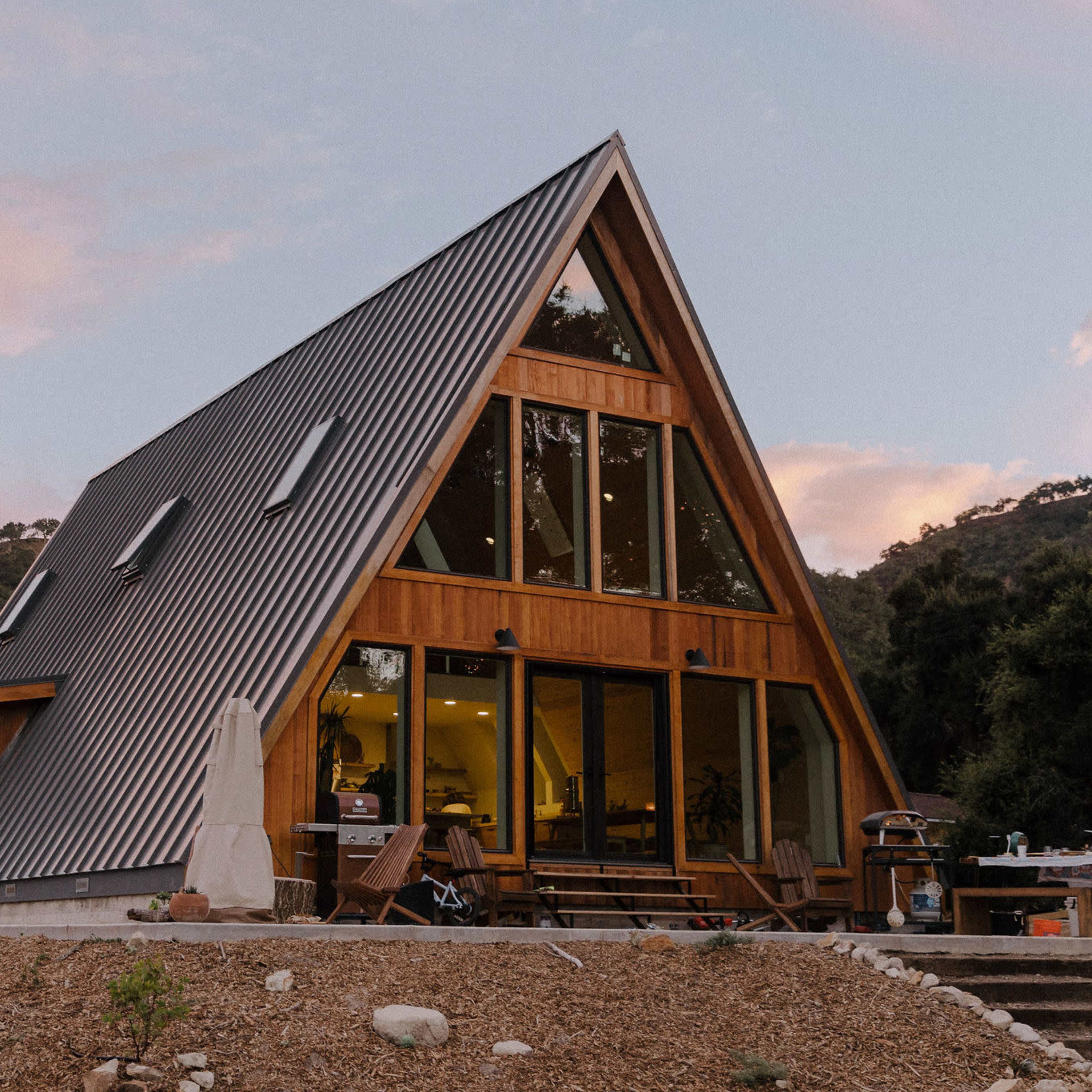 A modern A-frame house with large windows and a slanted metal roof is situated in a hilly landscape.