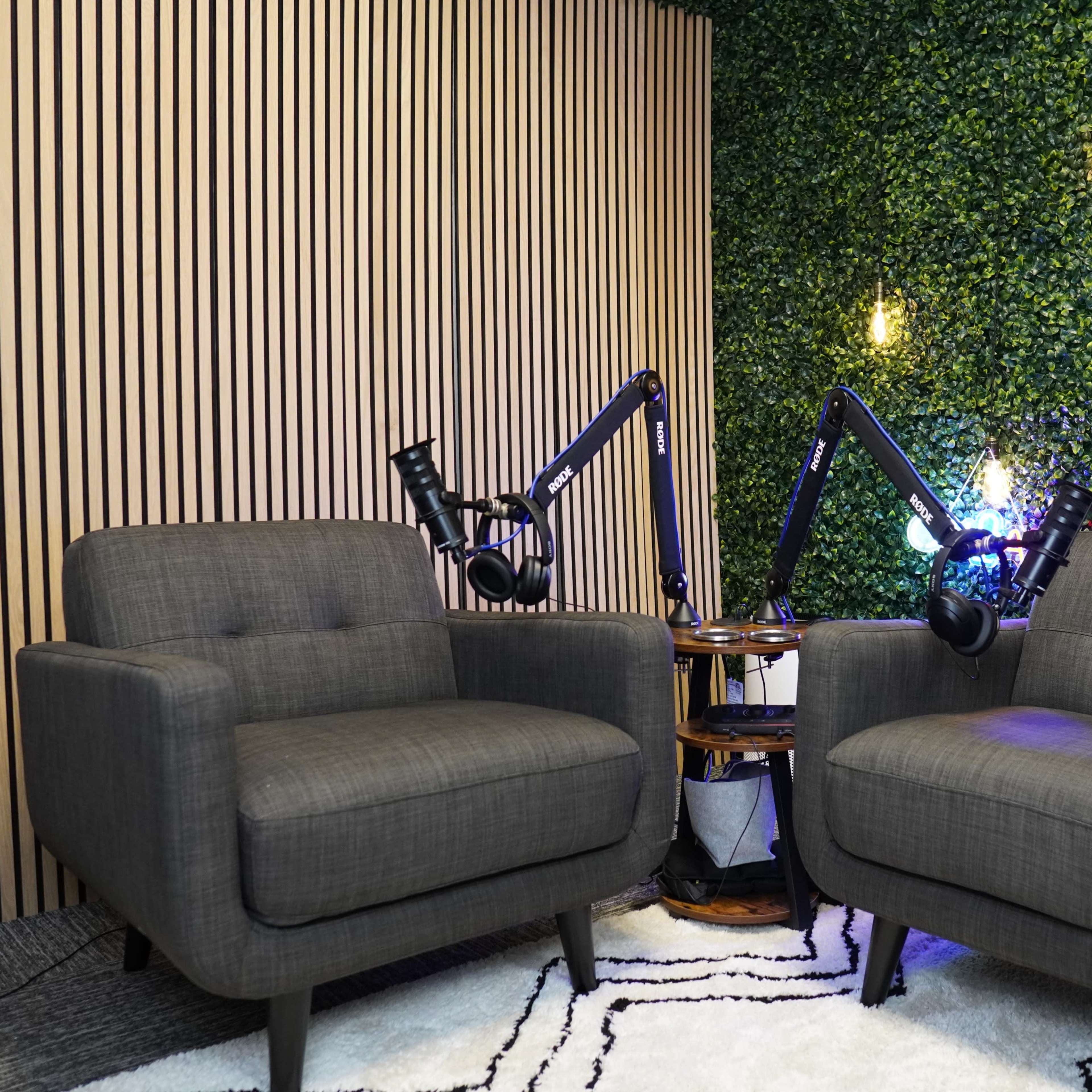 Two gray armchairs are positioned in a small studio with a green wall, equipped with adjustable microphone arms and hanging lights.