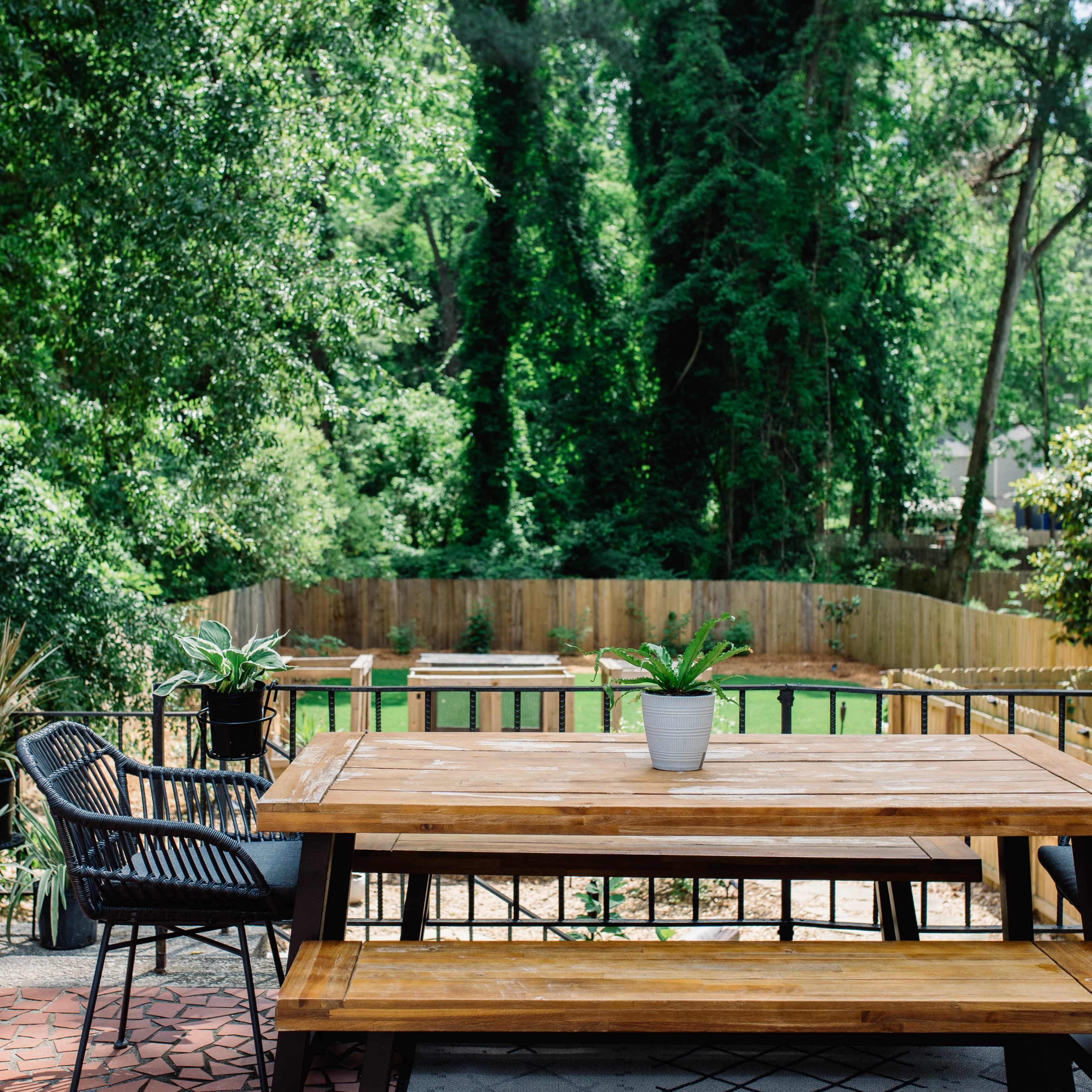 A wooden table with black wicker chairs is set on a patio overlooking a green lawn surrounded by trees and a wooden fence.