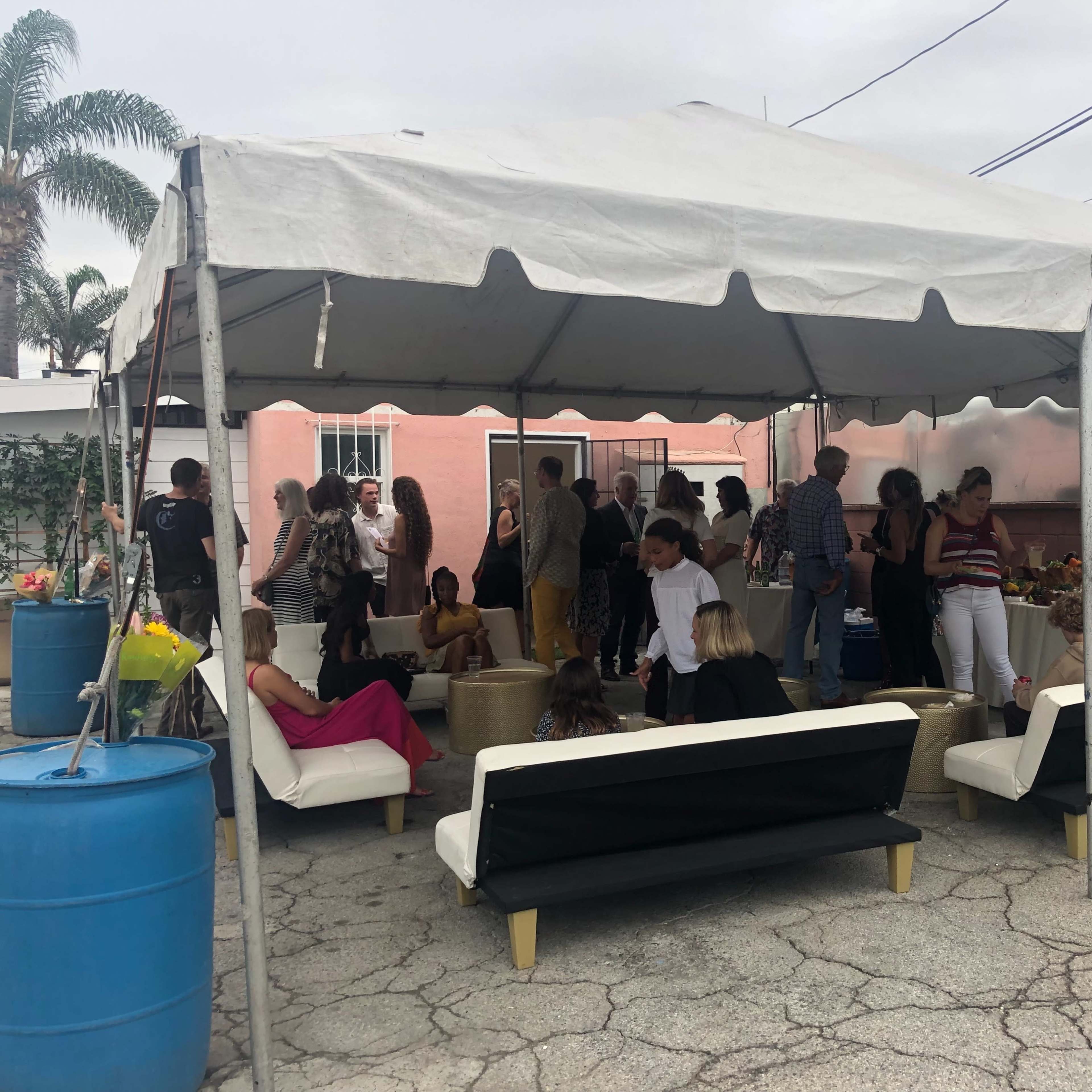 A large tent shelters a gathering of people socializing and seated on white sofas in an outdoor space.