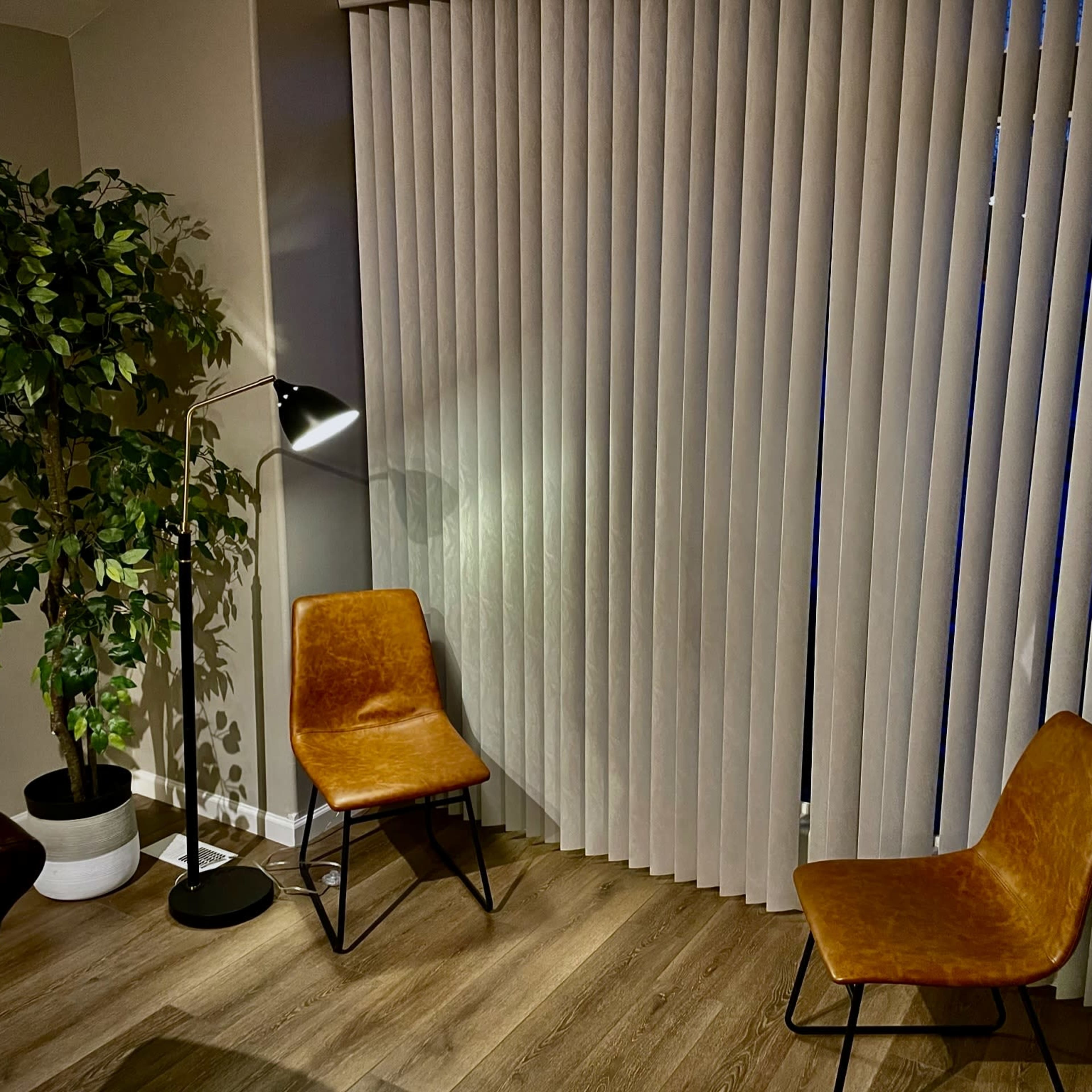 A corner of a room with two brown chairs, a floor lamp, and a potted plant next to vertically striped window coverings.