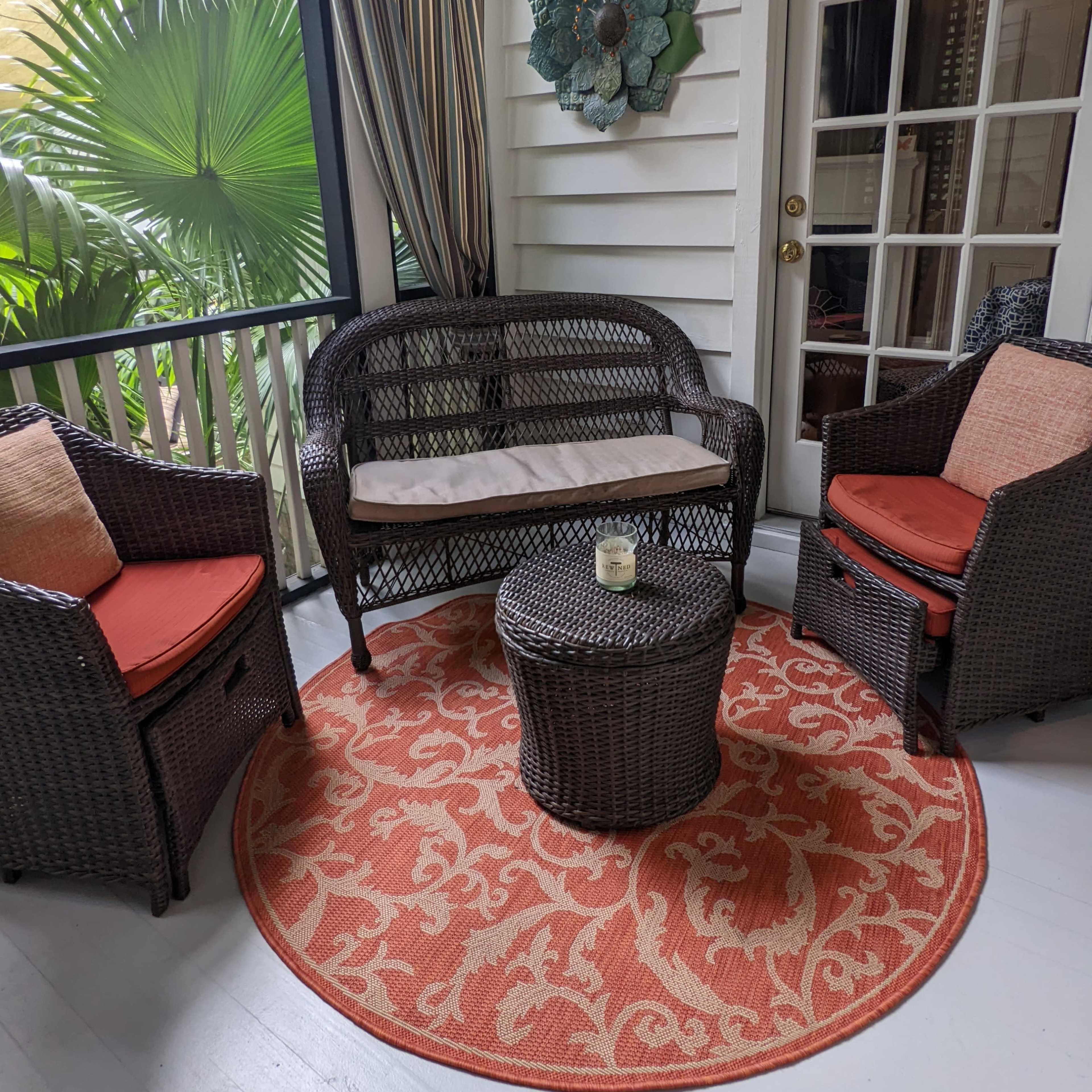 A small sitting area is set up on a porch with two wicker chairs and a round table, surrounded by plants and decorated with an orange patterned rug.