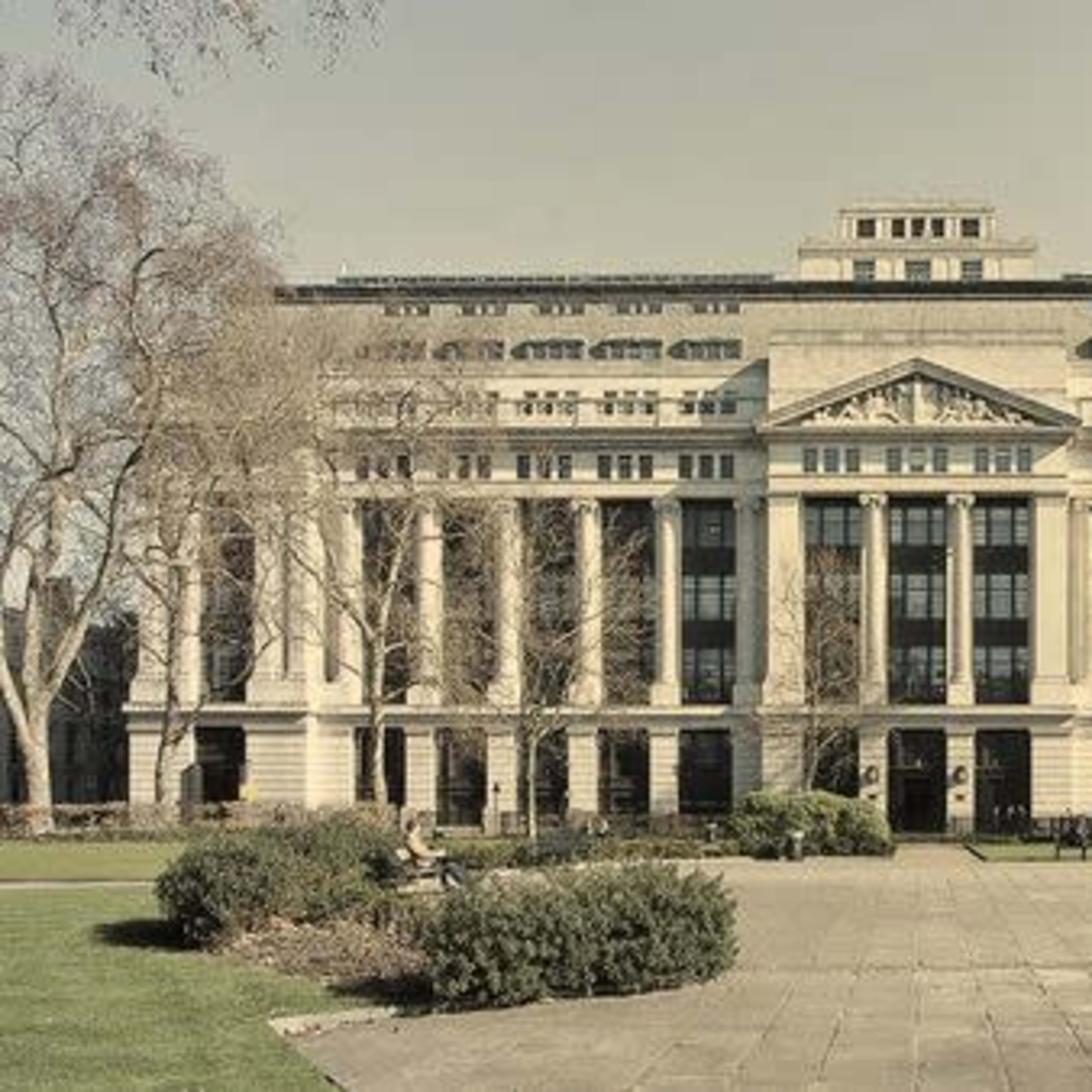 A large, classical-style building with columns and a symmetrical facade, surrounded by a park area with benches and trees.