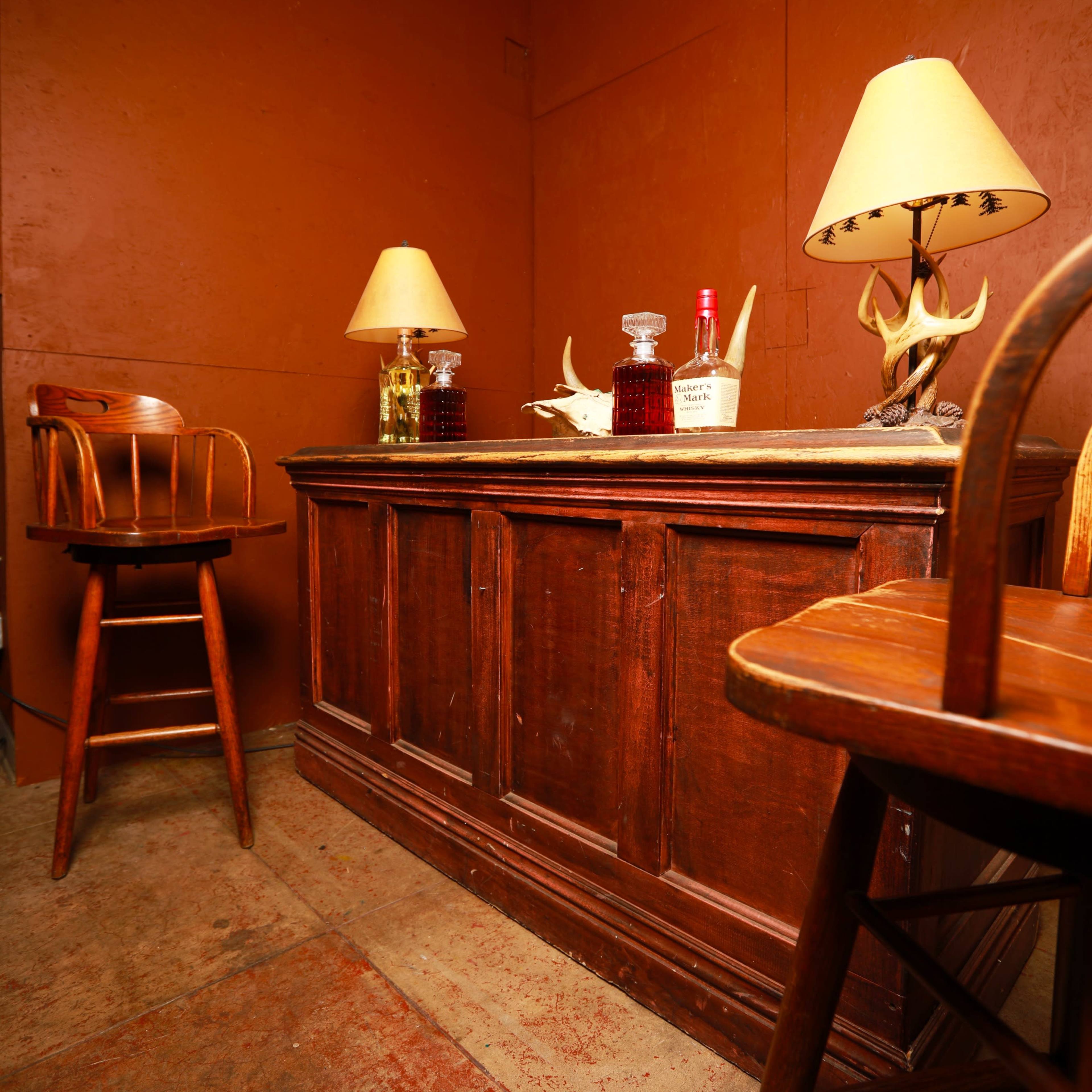A wooden bar is positioned against an orange wall, flanked by two lamps and a pair of wooden stools.