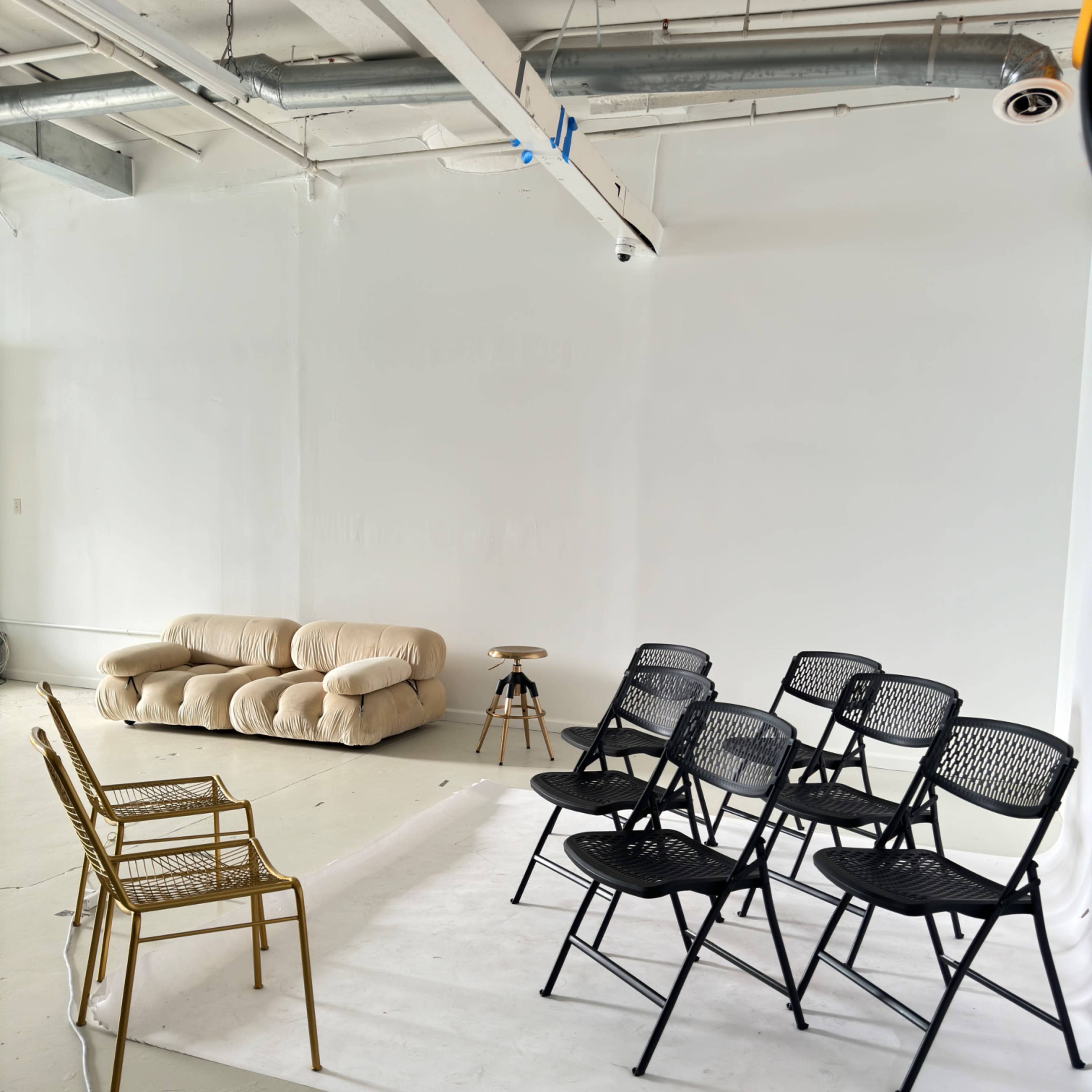 A beige couch is positioned against a white wall, facing a row of black folding chairs set up on a white backdrop in a spacious room.