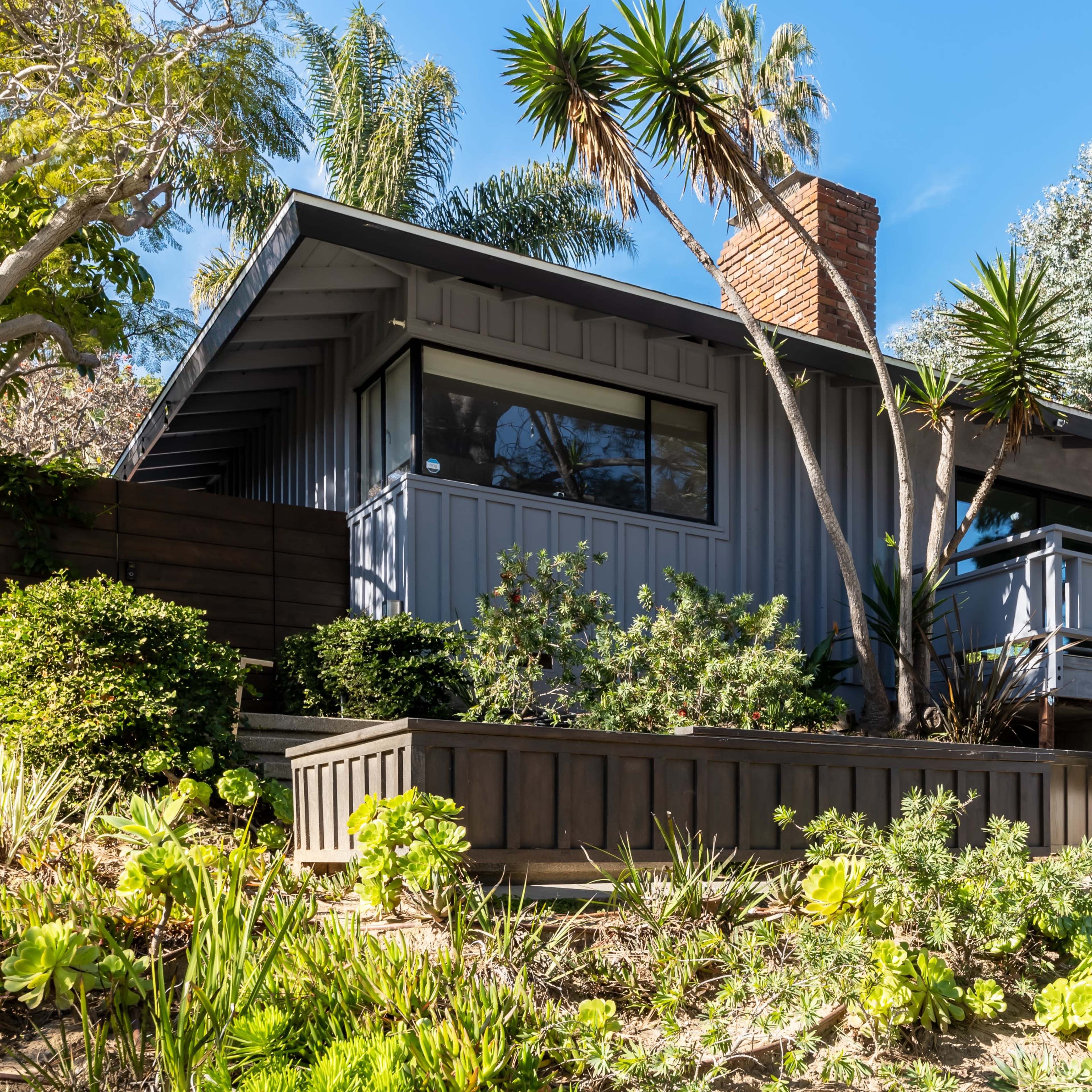 A modern house with a gray exterior and large windows is surrounded by lush greenery and landscaping on a hillside.