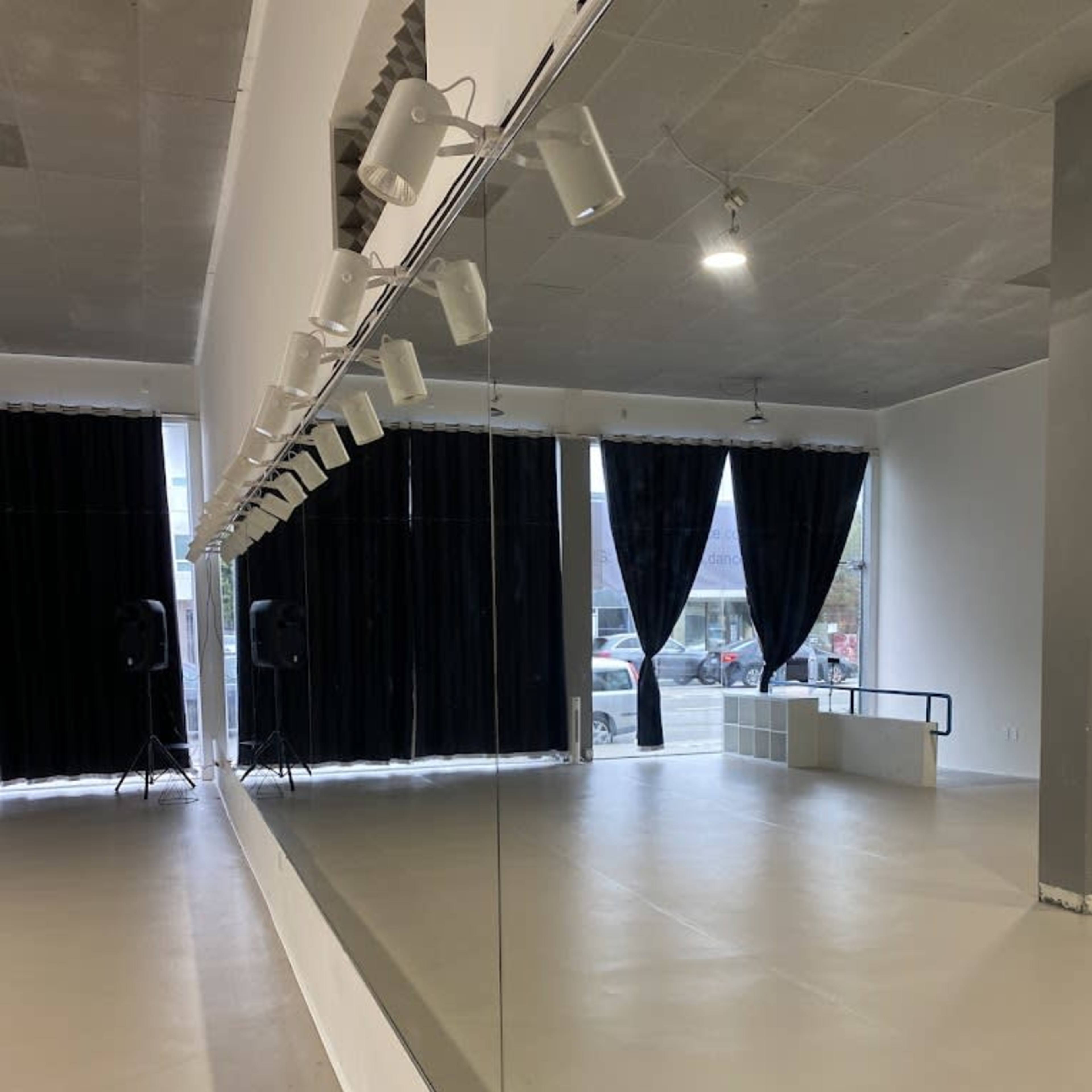 A dance studio features a large mirror along one wall, with black curtains covering the windows and speakers positioned in the corners.