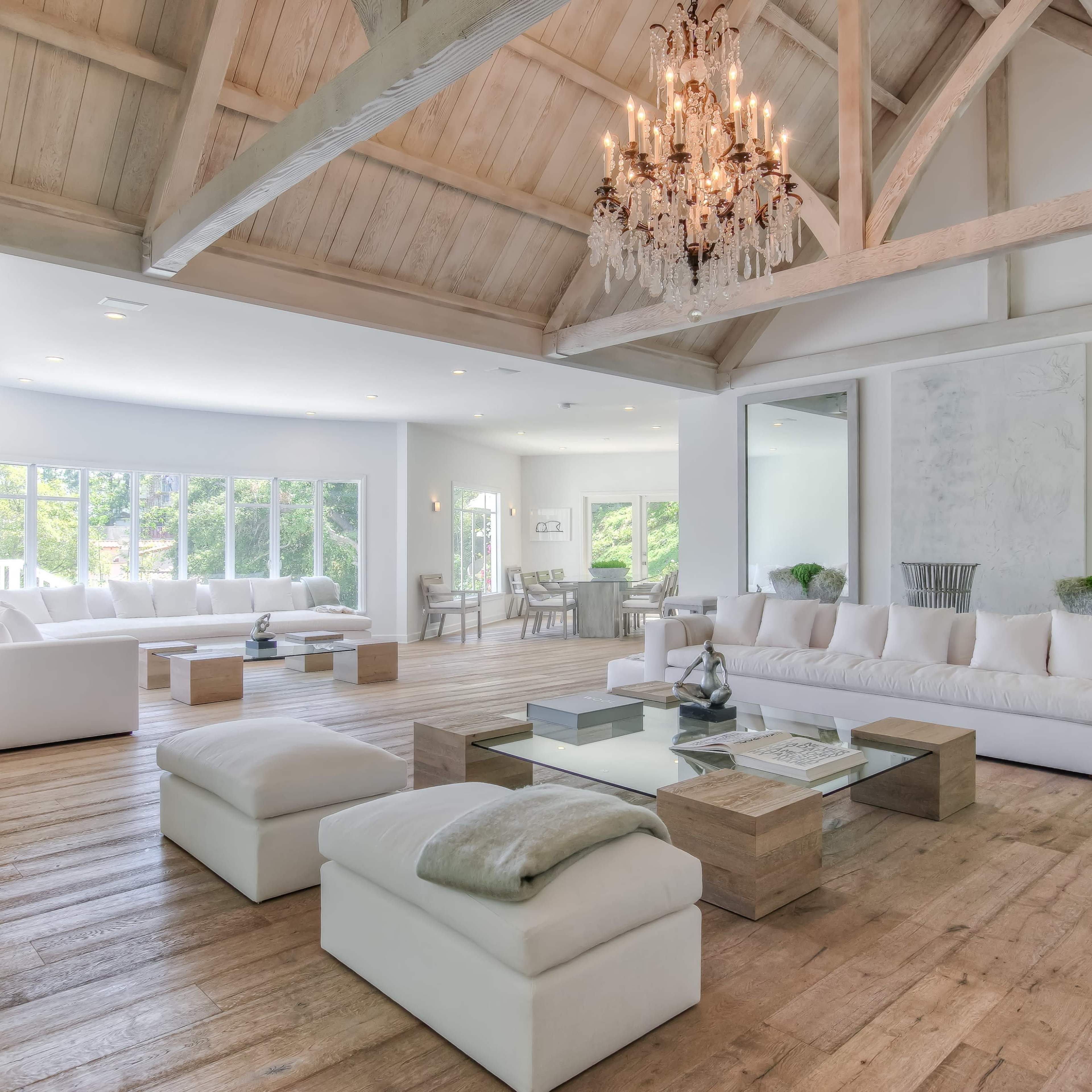 A spacious and bright living room features high wooden ceilings, large windows, and minimalist furniture arranged around a central coffee table.