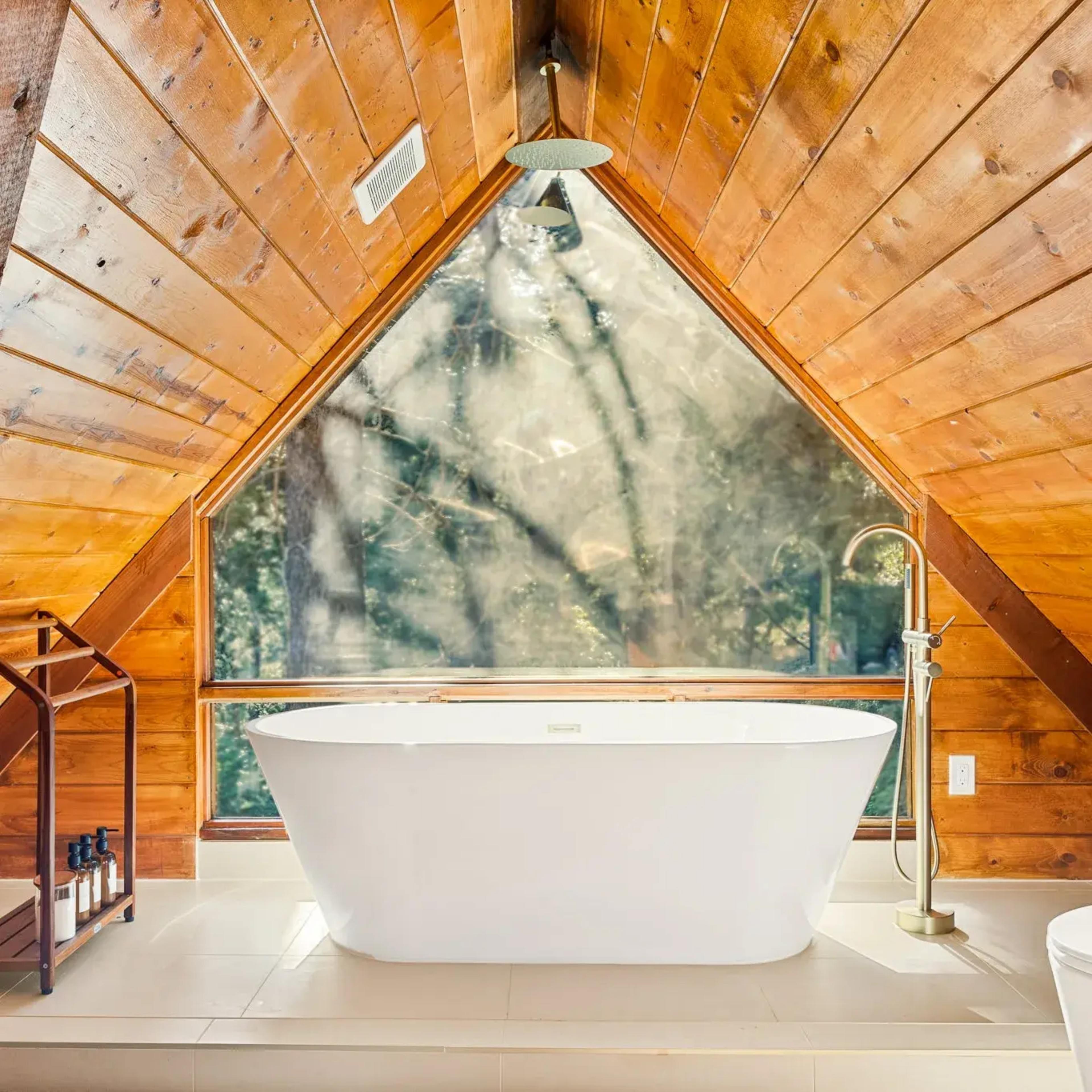 A modern, freestanding bathtub situated beneath a large triangular window in a wooden A-frame bathroom.