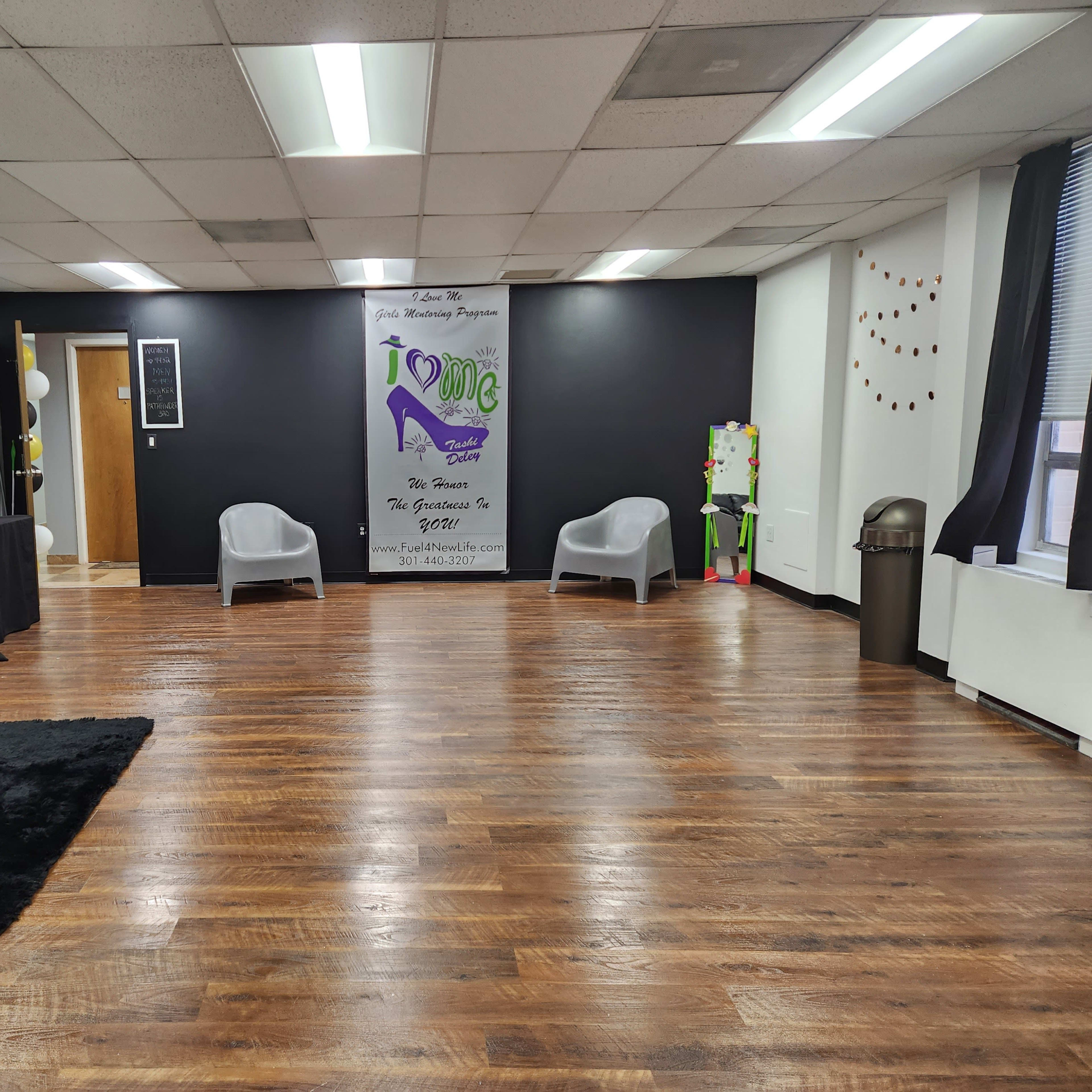 A spacious room with dark walls, hardwood flooring, and several seating areas, including two light gray chairs, a black couch, and a black table near the entrance.