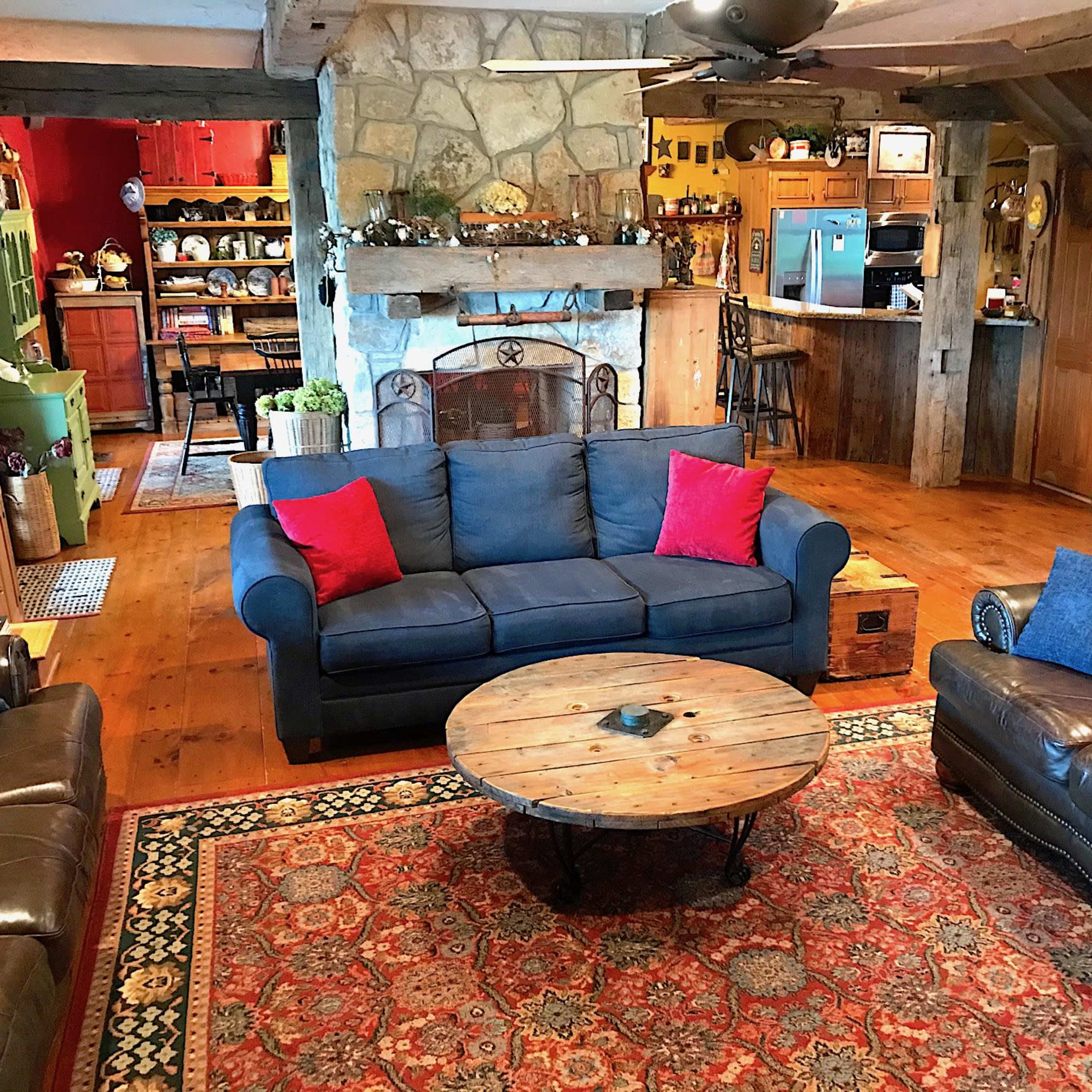 A cozy living room features a blue sofa with red cushions, a round wooden coffee table, and a stone fireplace, surrounded by wooden beams and rustic decor.