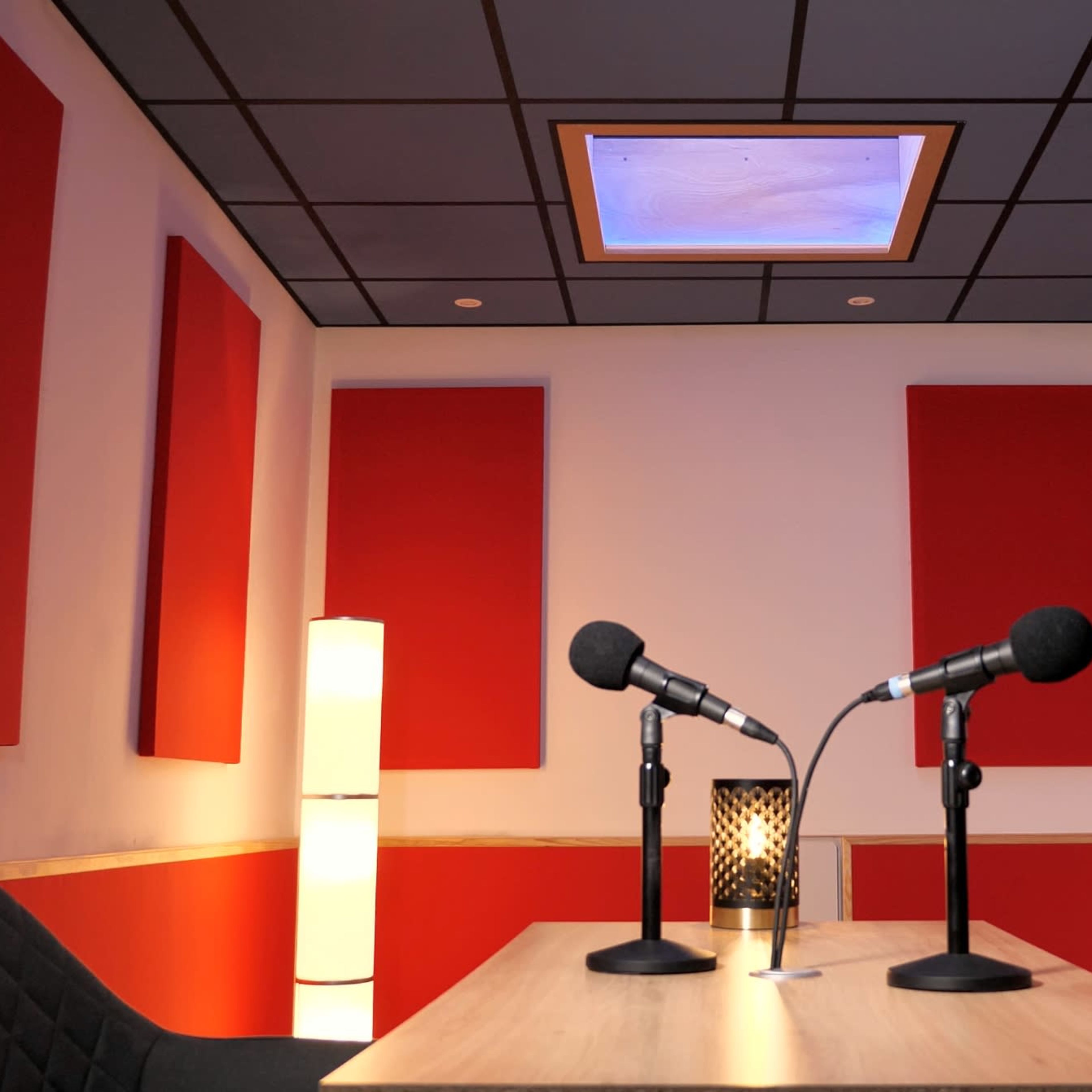 A soundproof recording studio featuring two microphones on a table, red acoustic panels on the walls, and a lamp in the corner.