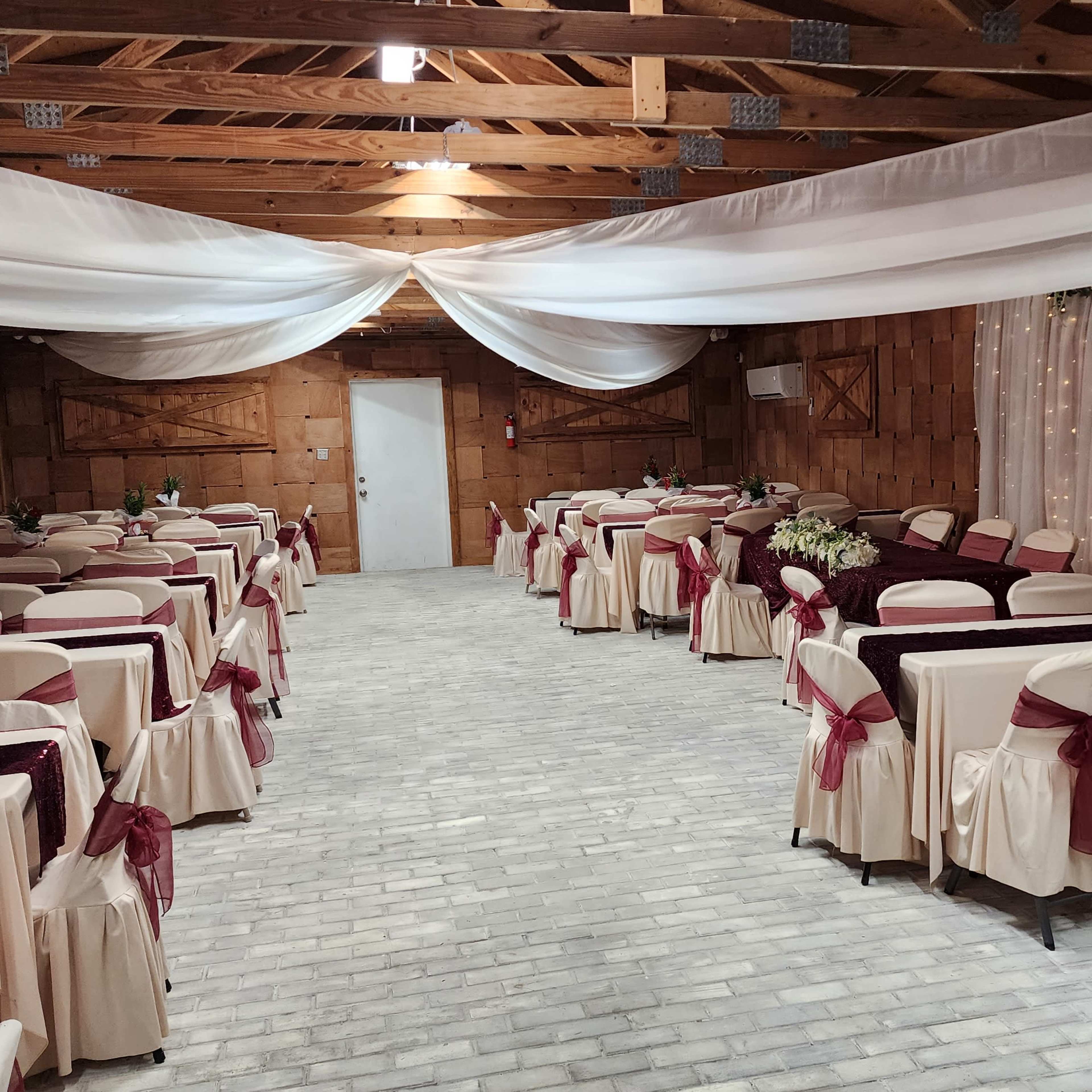A banquet hall set up with rows of tables covered in beige tablecloths and burgundy sashes, under draped white fabric and soft lighting.