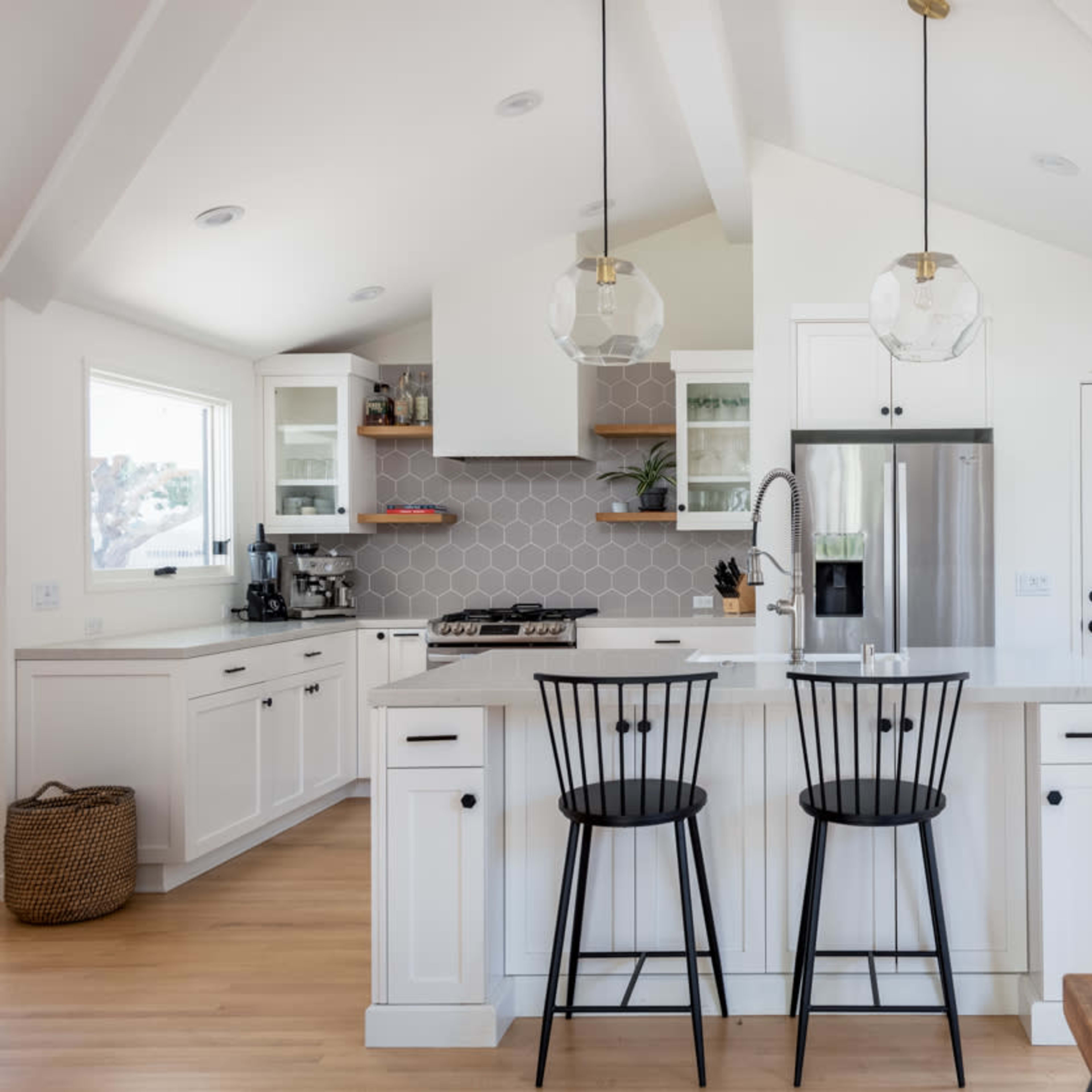A modern kitchen features white cabinetry, a central island with black bar stools, and large windows allowing natural light to enter.