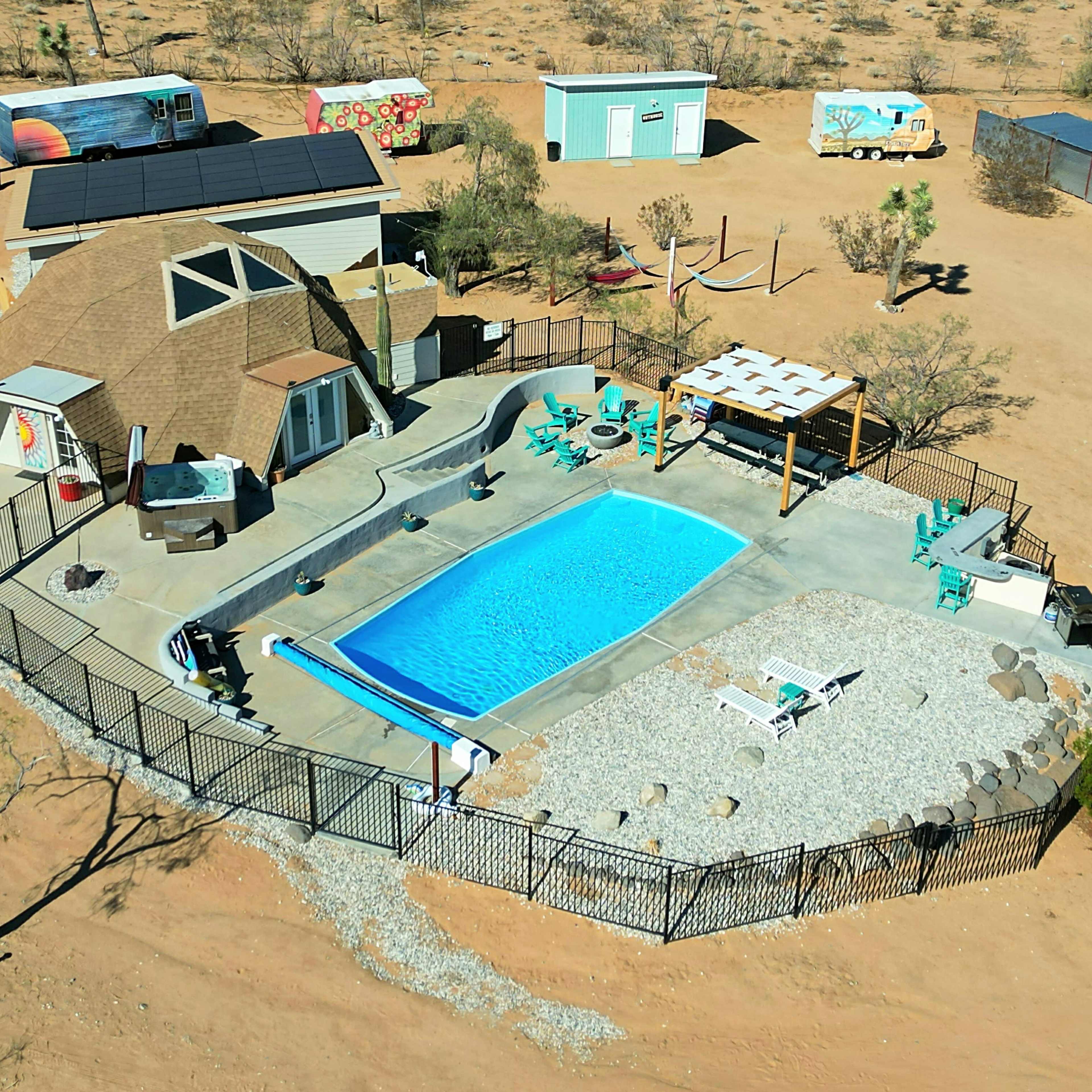 A desert property featuring a dome-shaped house, a swimming pool surrounded by a fence, a shaded seating area, and various outdoor structures.