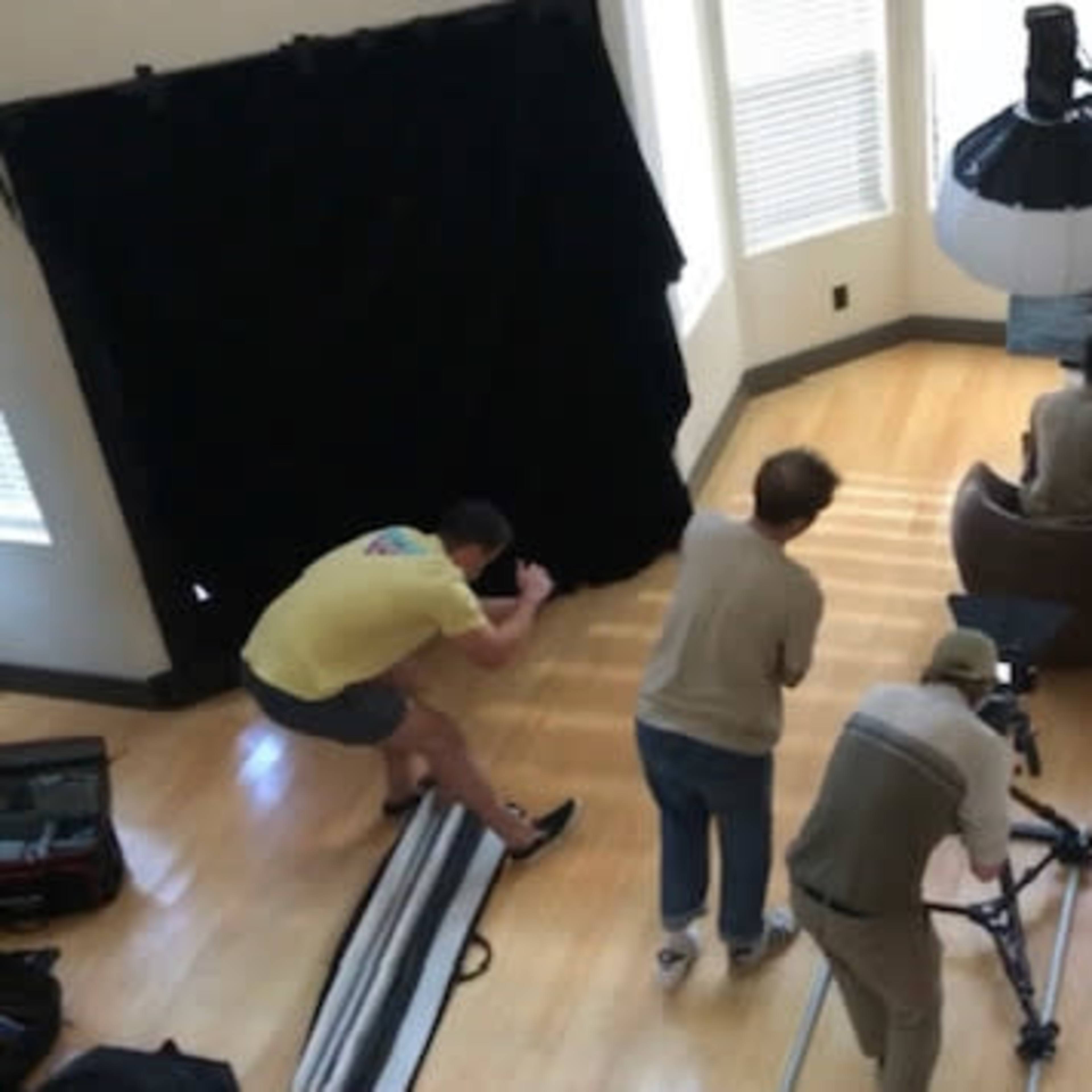A photography crew sets up a shoot in a well-lit room featuring a model seated in a large round chair, with various equipment positioned around them.