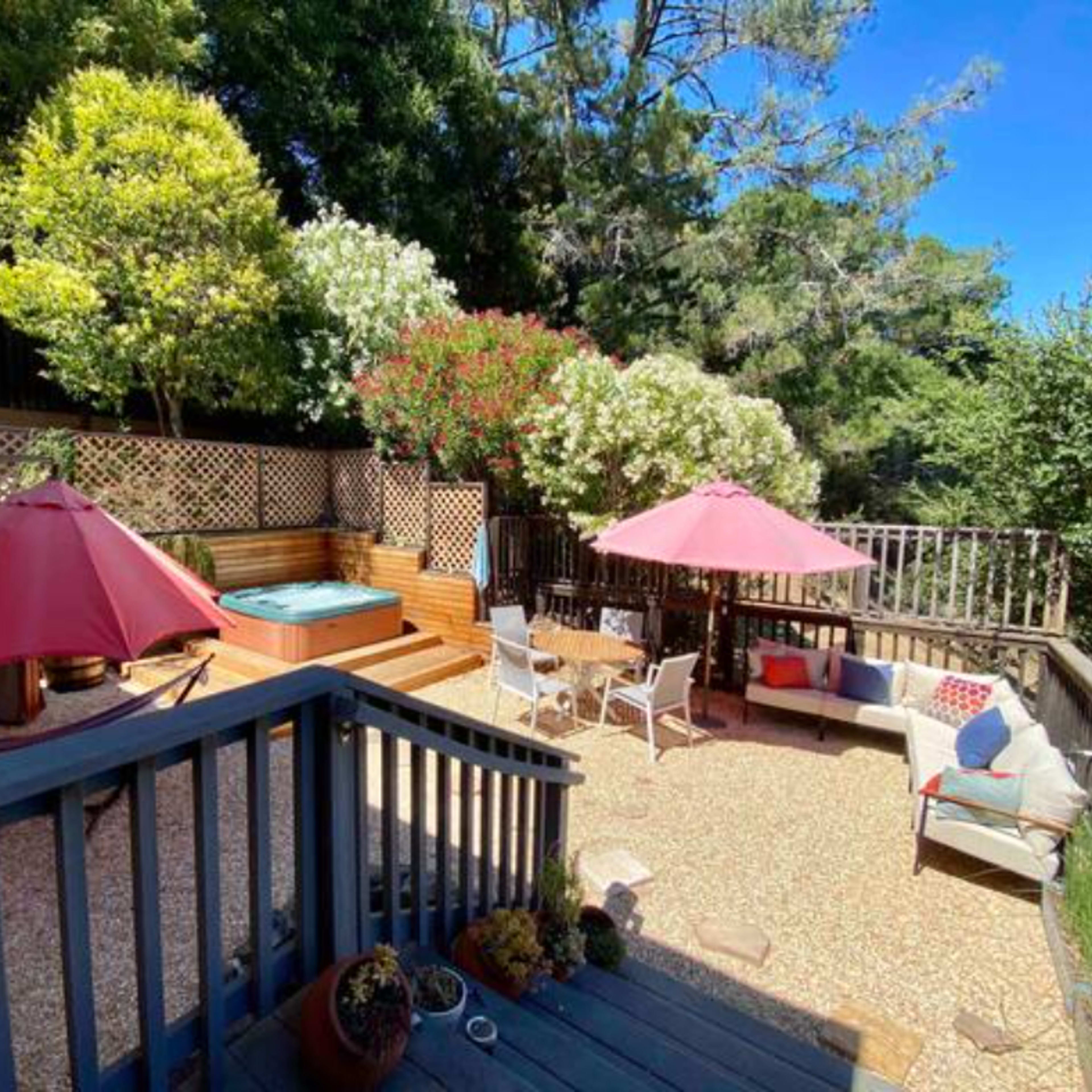 A backyard patio with a hot tub, surrounded by wooden decking, flowering plants, two pink umbrellas, and outdoor seating.