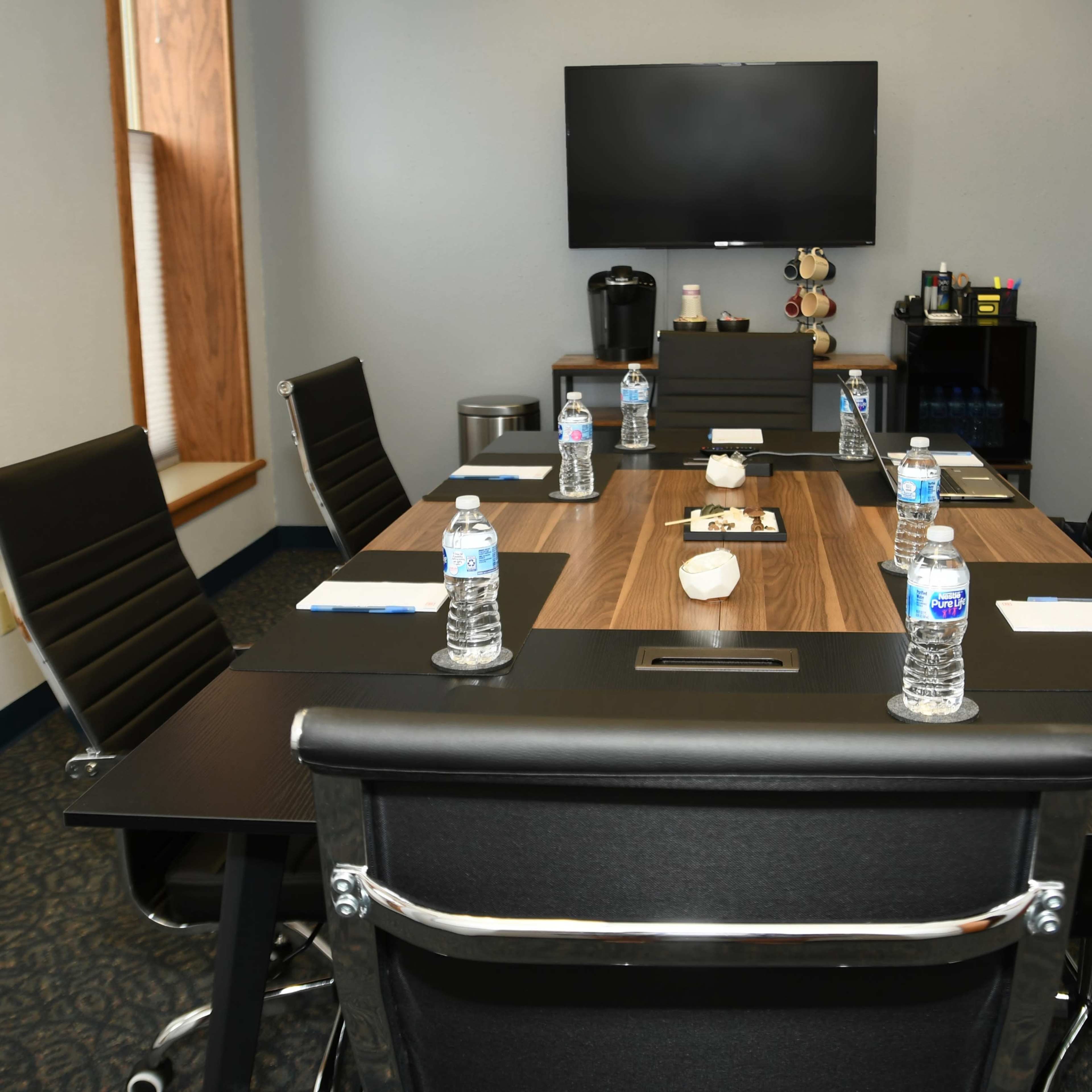 A conference room is set up with a long table surrounded by six chairs, each featuring a water bottle, utensils, and notepads, while a television is mounted on the wall.