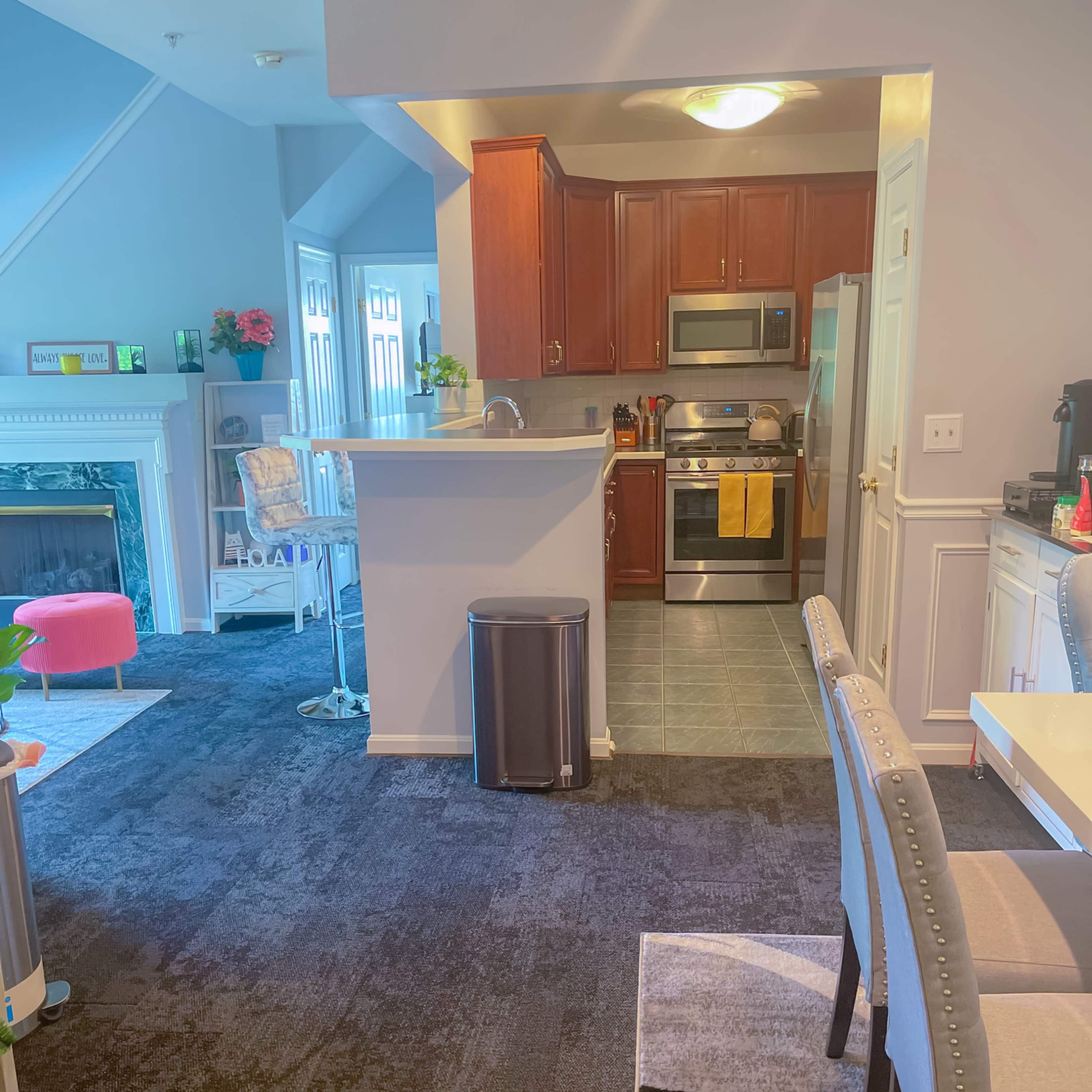 A modern kitchen area with wood cabinets, stainless steel appliances, and a dining space adjacent to it, featuring a table and chairs on a dark carpet.
