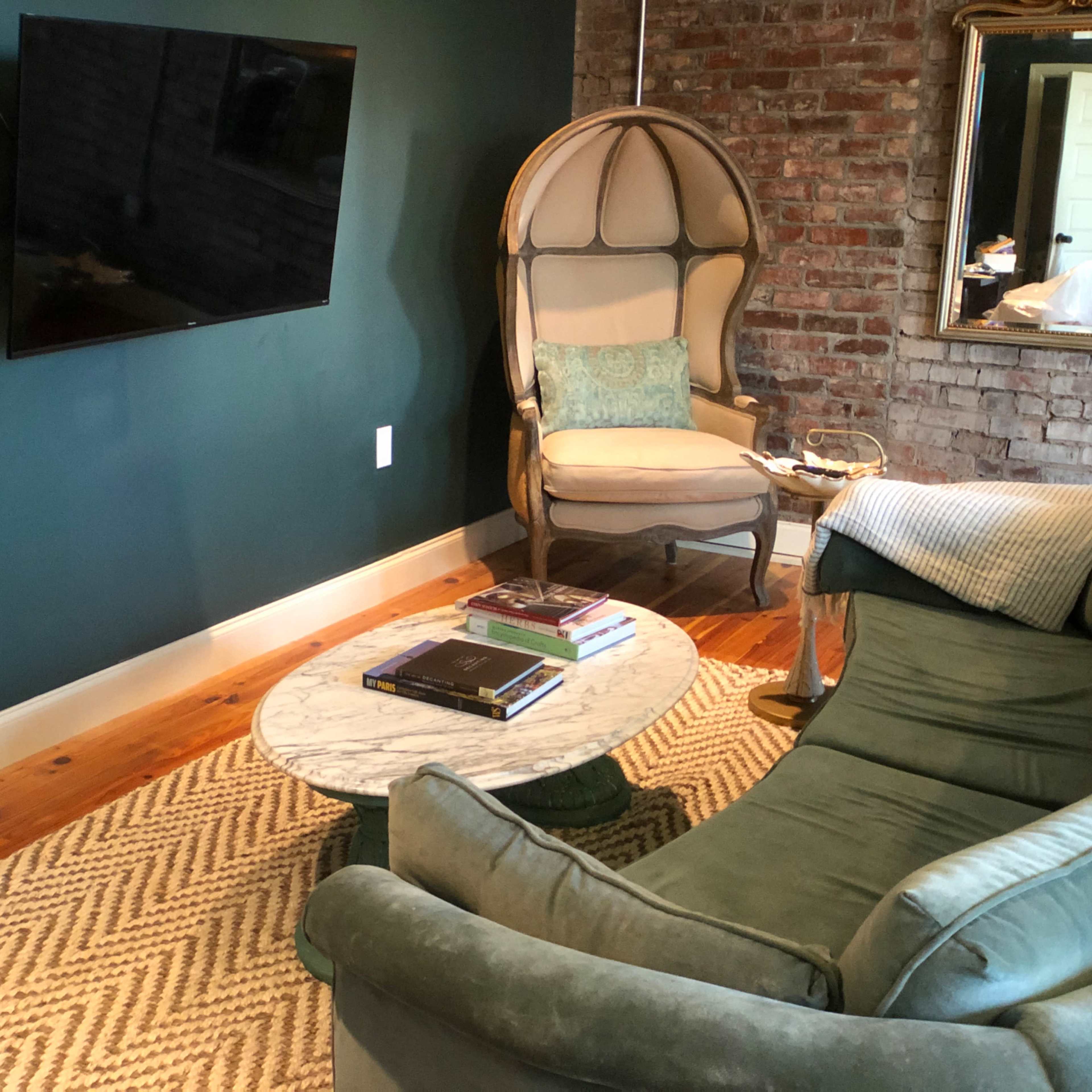 A cozy living space features a green wall, a mounted TV, a vintage chair, and a marble coffee table surrounded by a sofa and a woven rug.