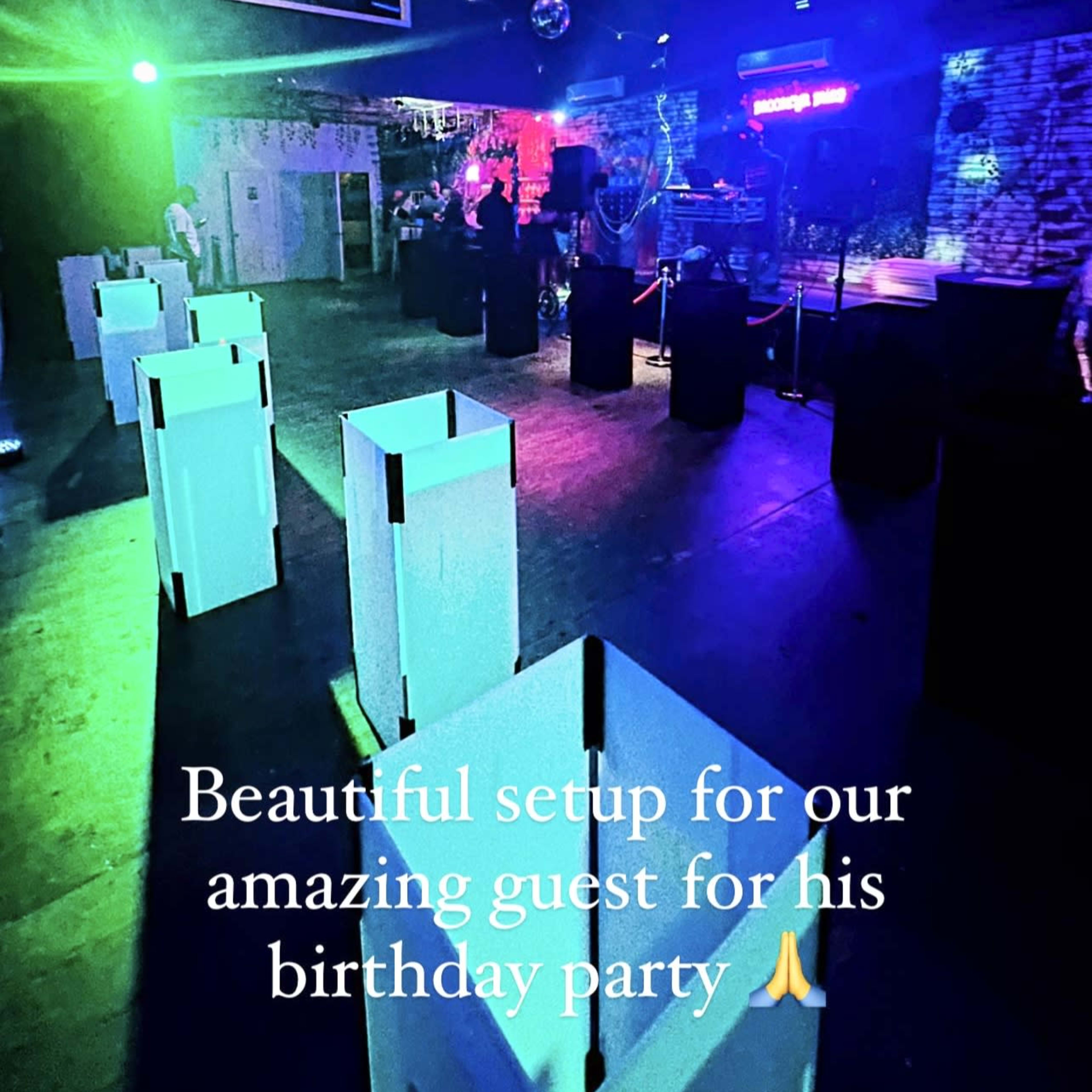 A dimly lit venue is arranged for a birthday party, featuring colorful lighting and several white tables.