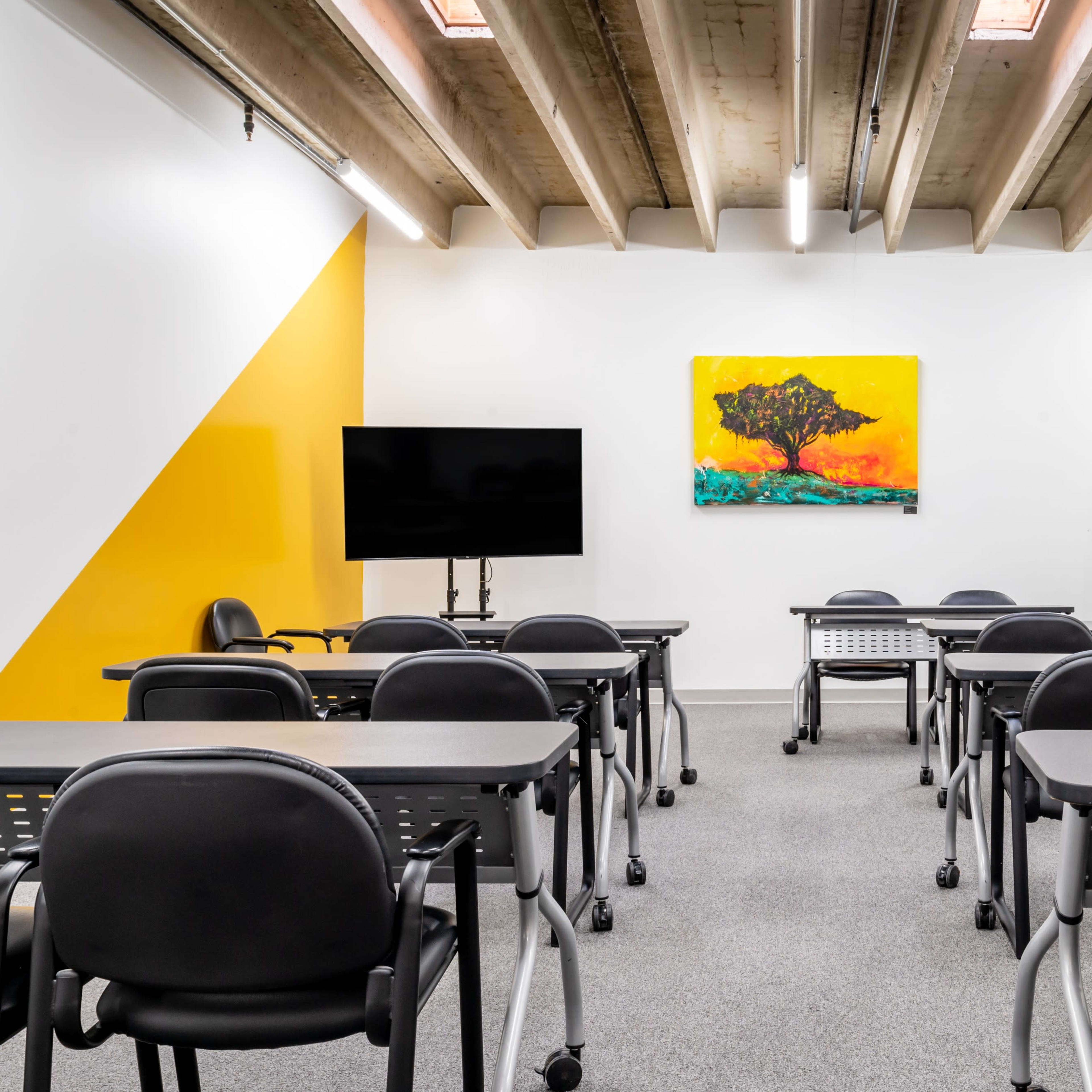 A classroom is arranged with black desks and chairs facing a large wall-mounted screen and a colorful artwork of a tree.